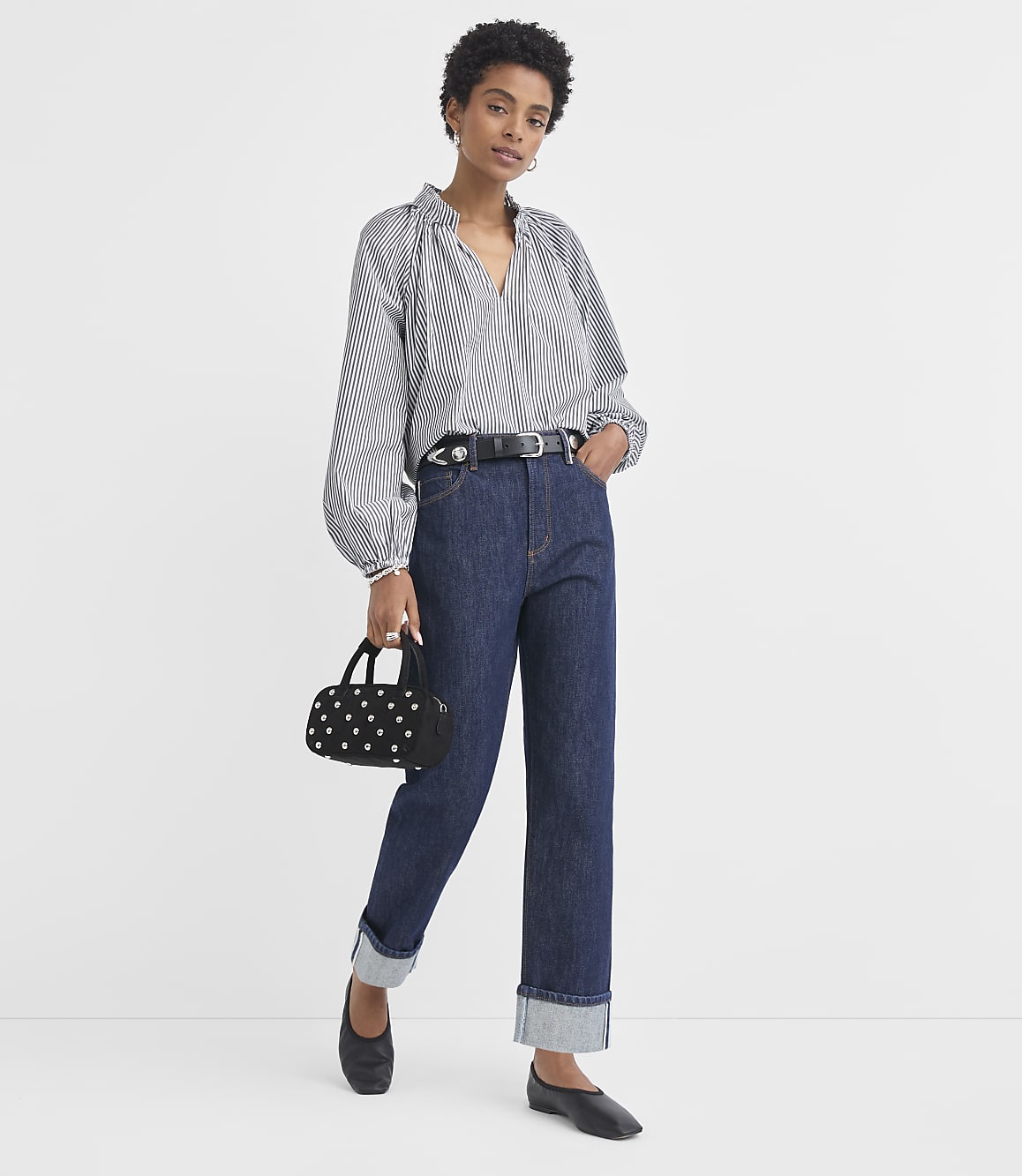 A woman in a striped blouse and dark jeans stands against a plain white background, holding a small black purse.