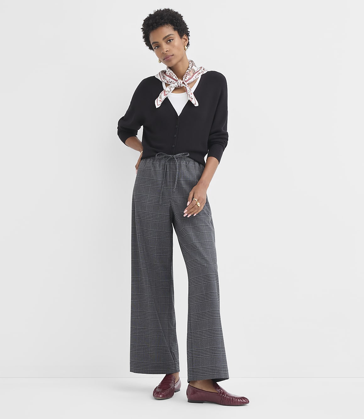 A woman in a black cardigan and plaid pants stands against a plain white background.