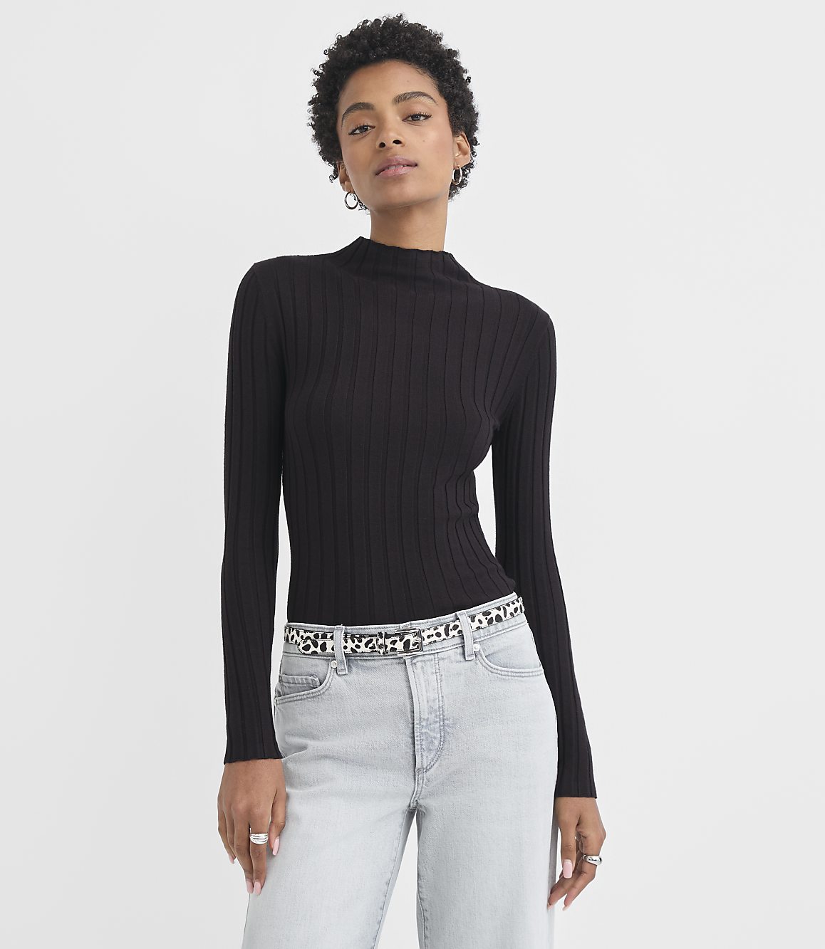 A woman in a black ribbed turtleneck and light wash jeans stands against a plain white background.