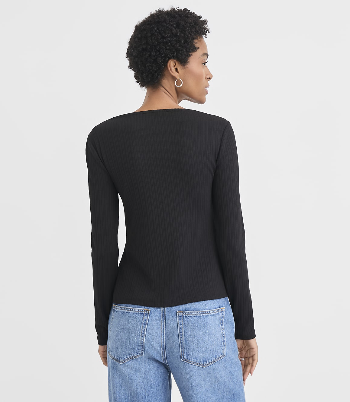 A woman in a black ribbed long-sleeved shirt and blue jeans stands against a plain white background.