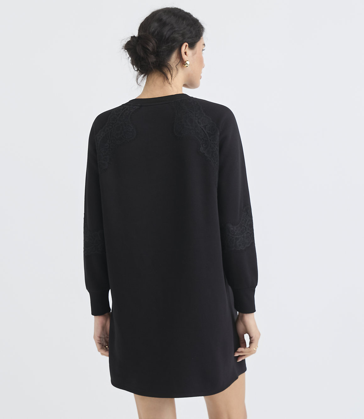 A woman in a black dress with lace detailing on the shoulders and sleeves stands against a plain white background.