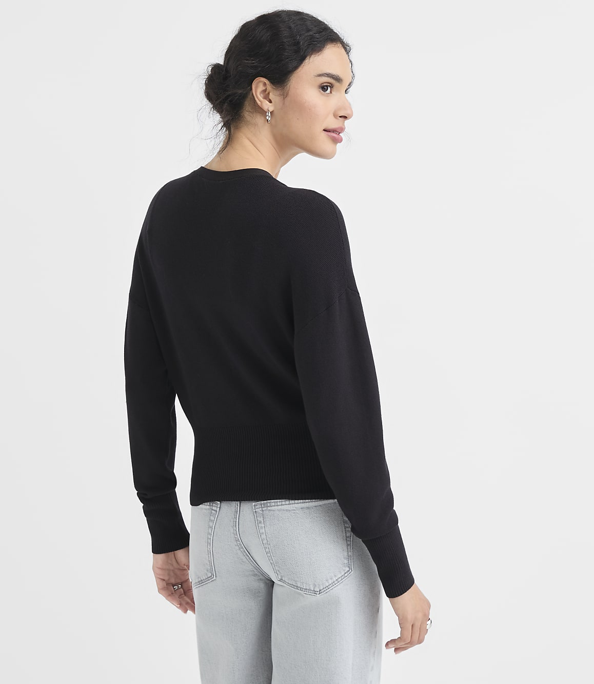 A woman in a black sweater and light-wash jeans stands against a plain white background.