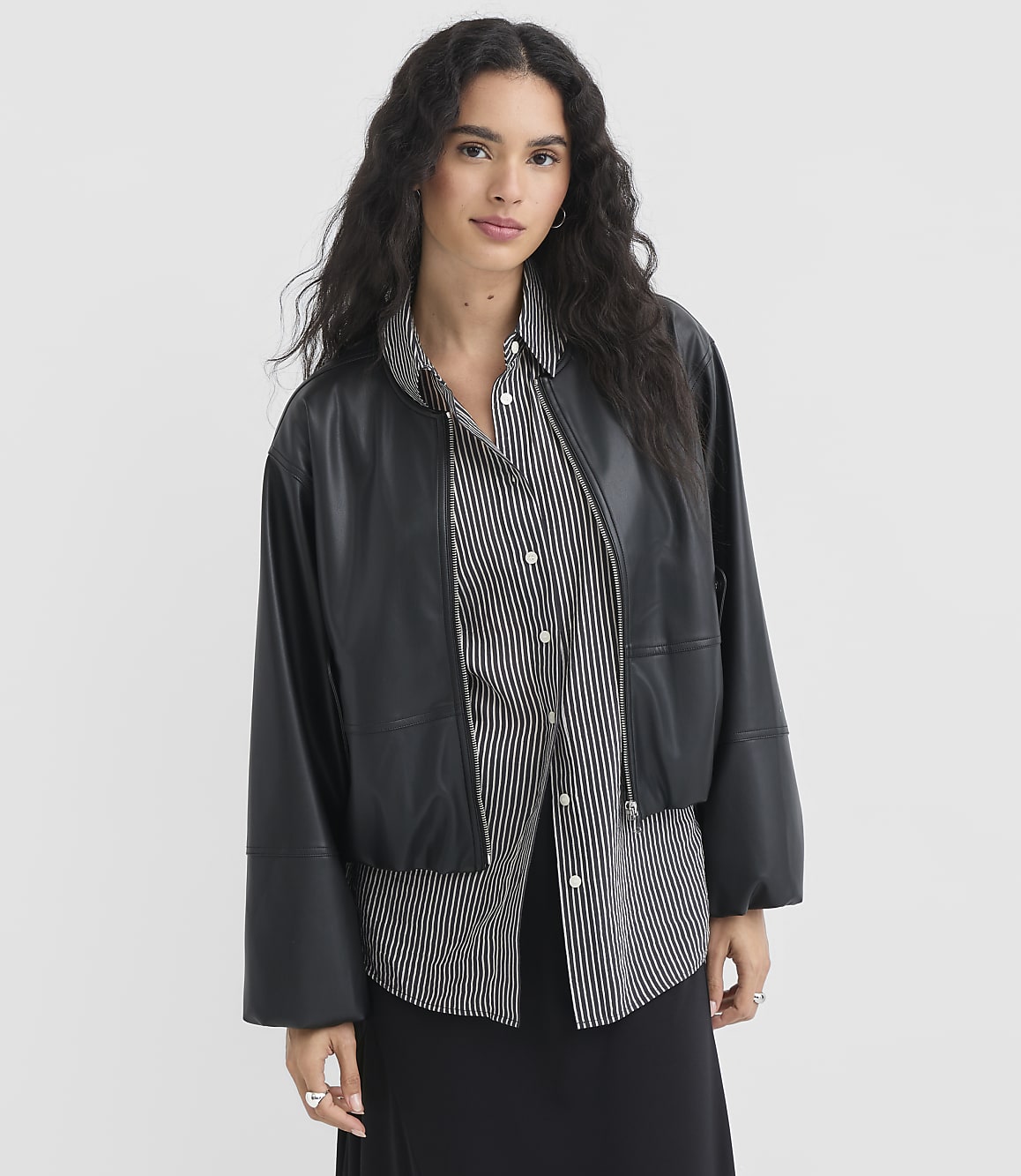 A woman in a black leather jacket and striped shirt stands against a plain white background.