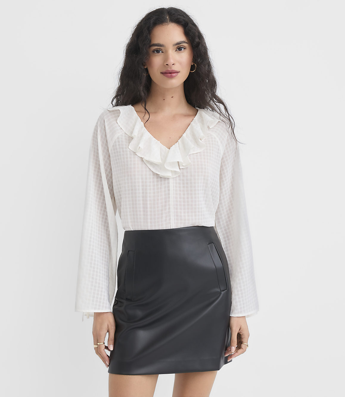 A woman in a white ruffled blouse and black leather skirt stands against a plain white background.
