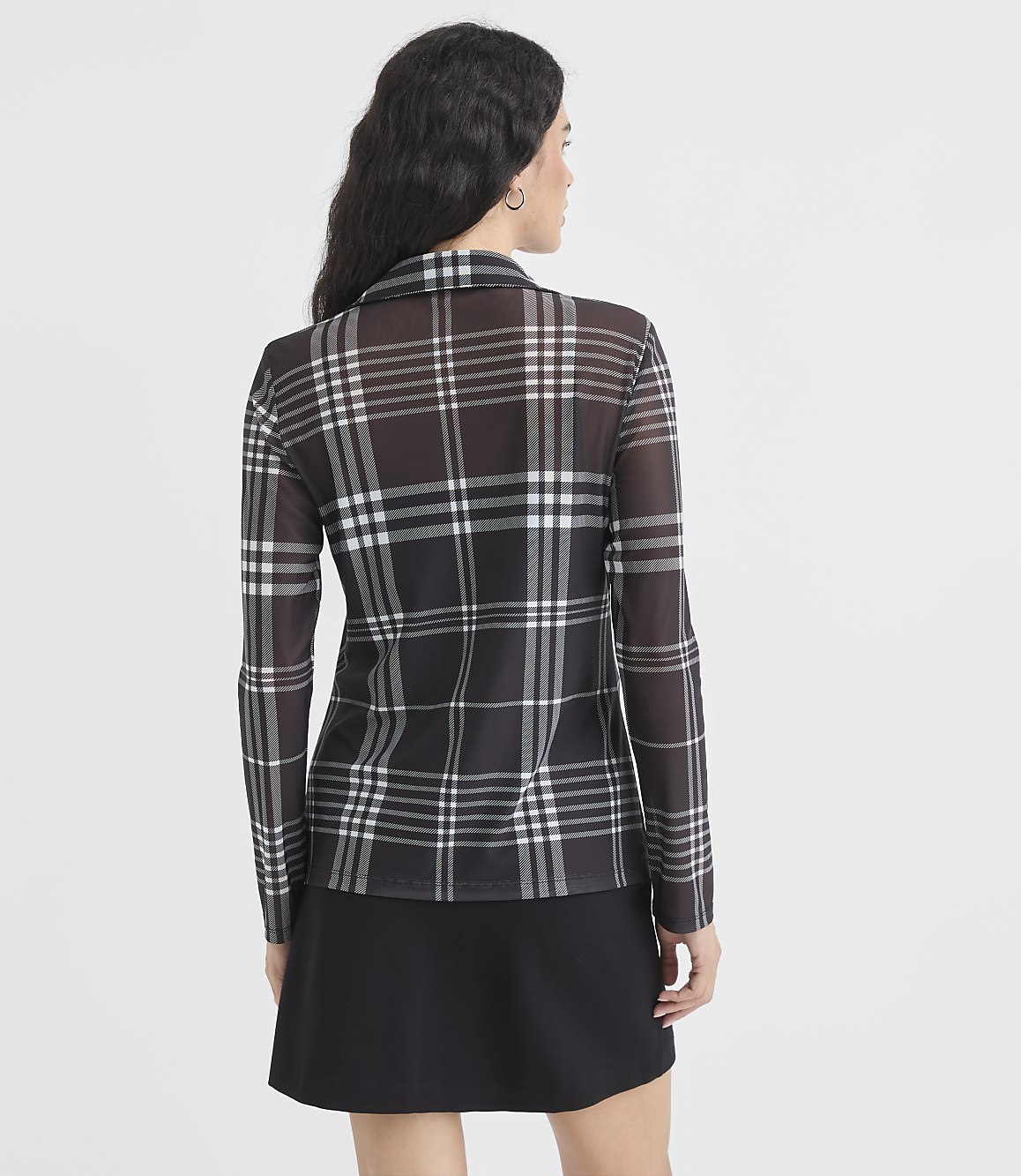 A woman in a sheer plaid top and black skirt stands against a plain white background.