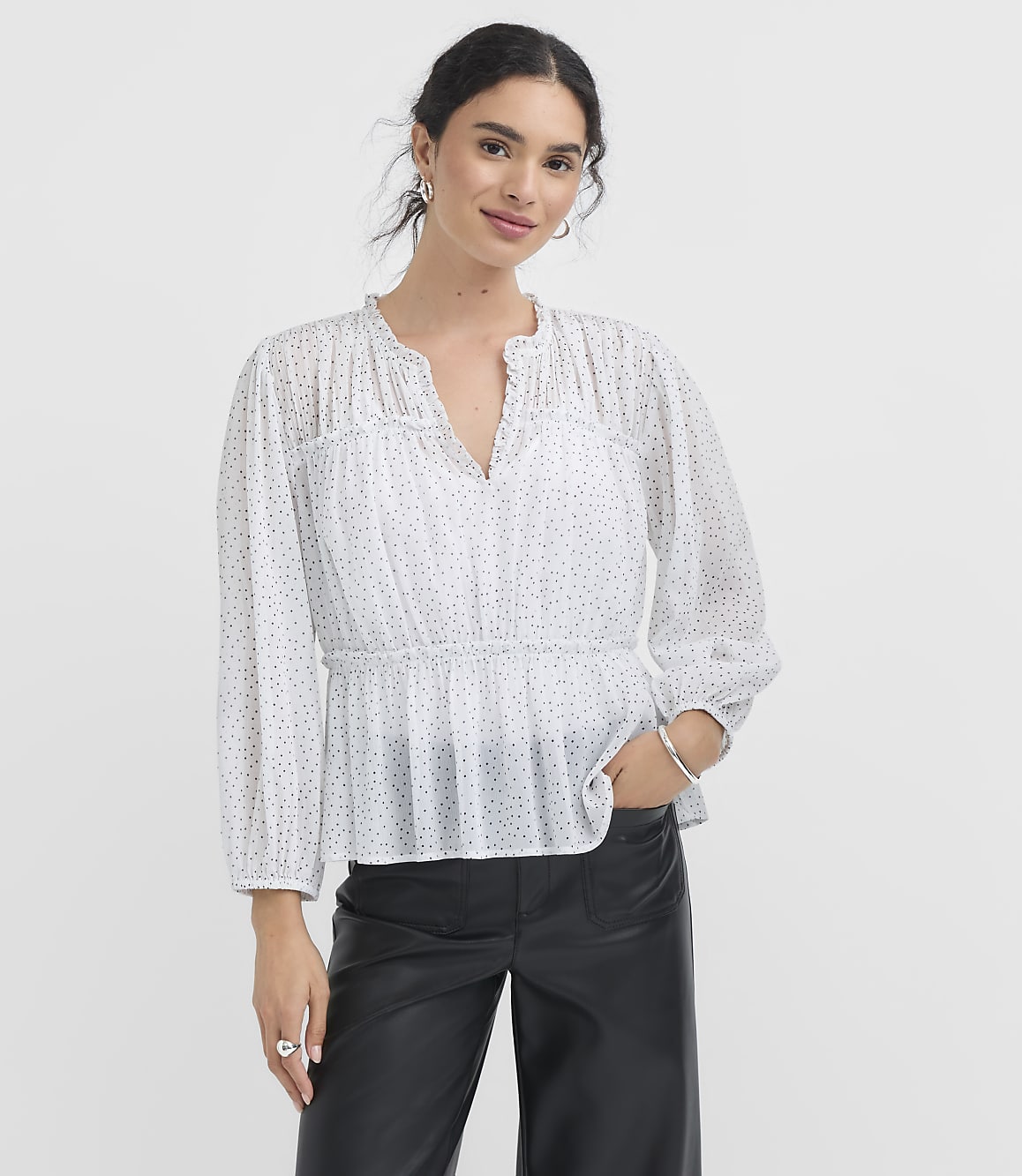 A woman in a white polka dot blouse and black pants stands against a plain white background.