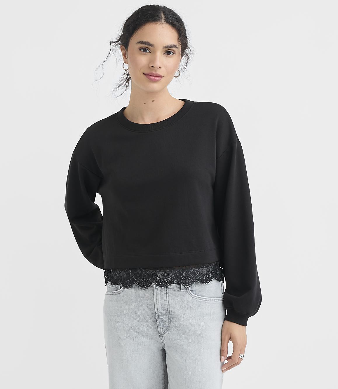 A young woman in a black cropped sweatshirt with lace trim and light wash jeans stands against a plain white background.