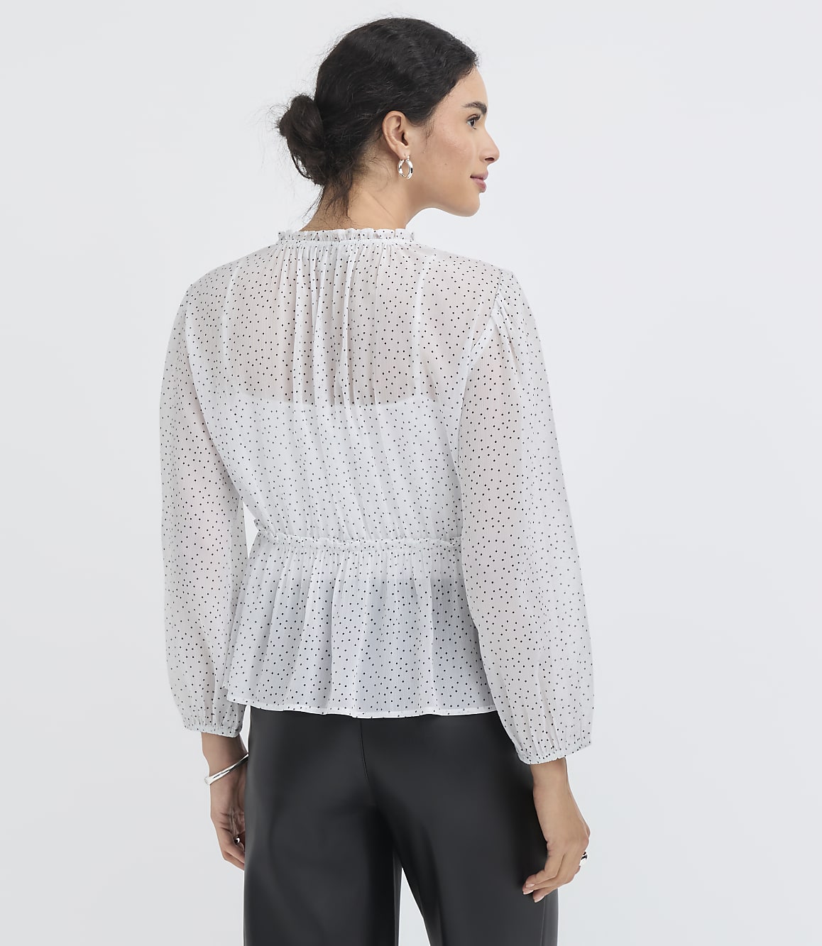 A woman in a sheer polka-dot peplum blouse and black pants stands against a plain white background.