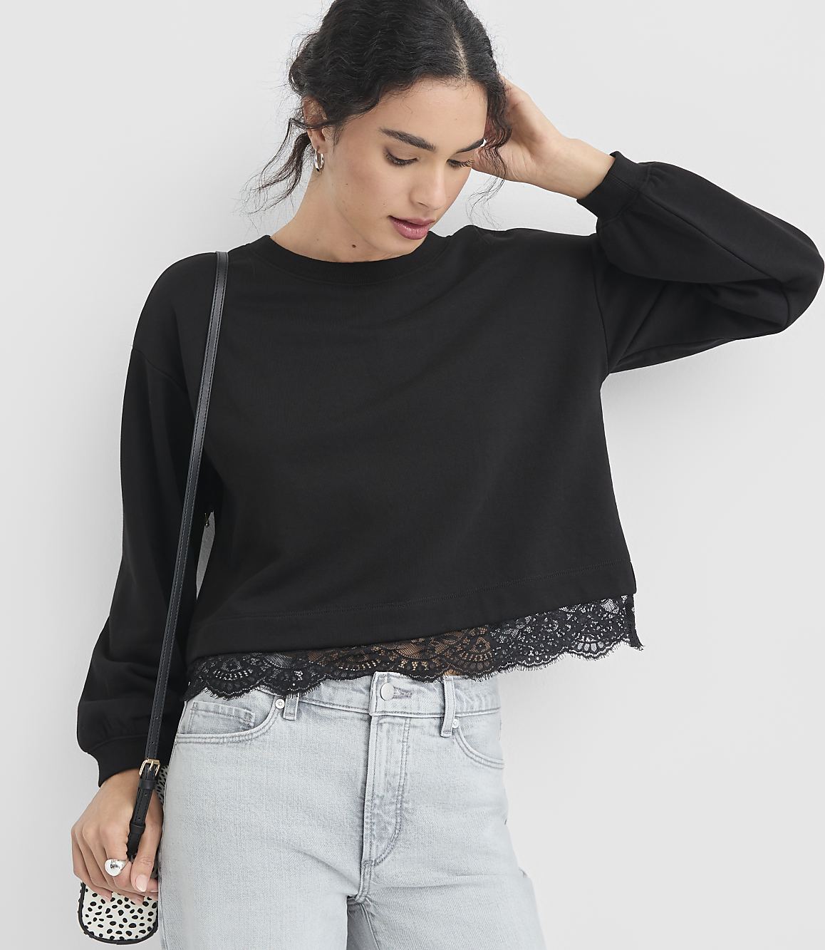 A woman in a black lace-trimmed sweatshirt and light-wash jeans stands against a plain white background, with a small patterned crossbody bag.