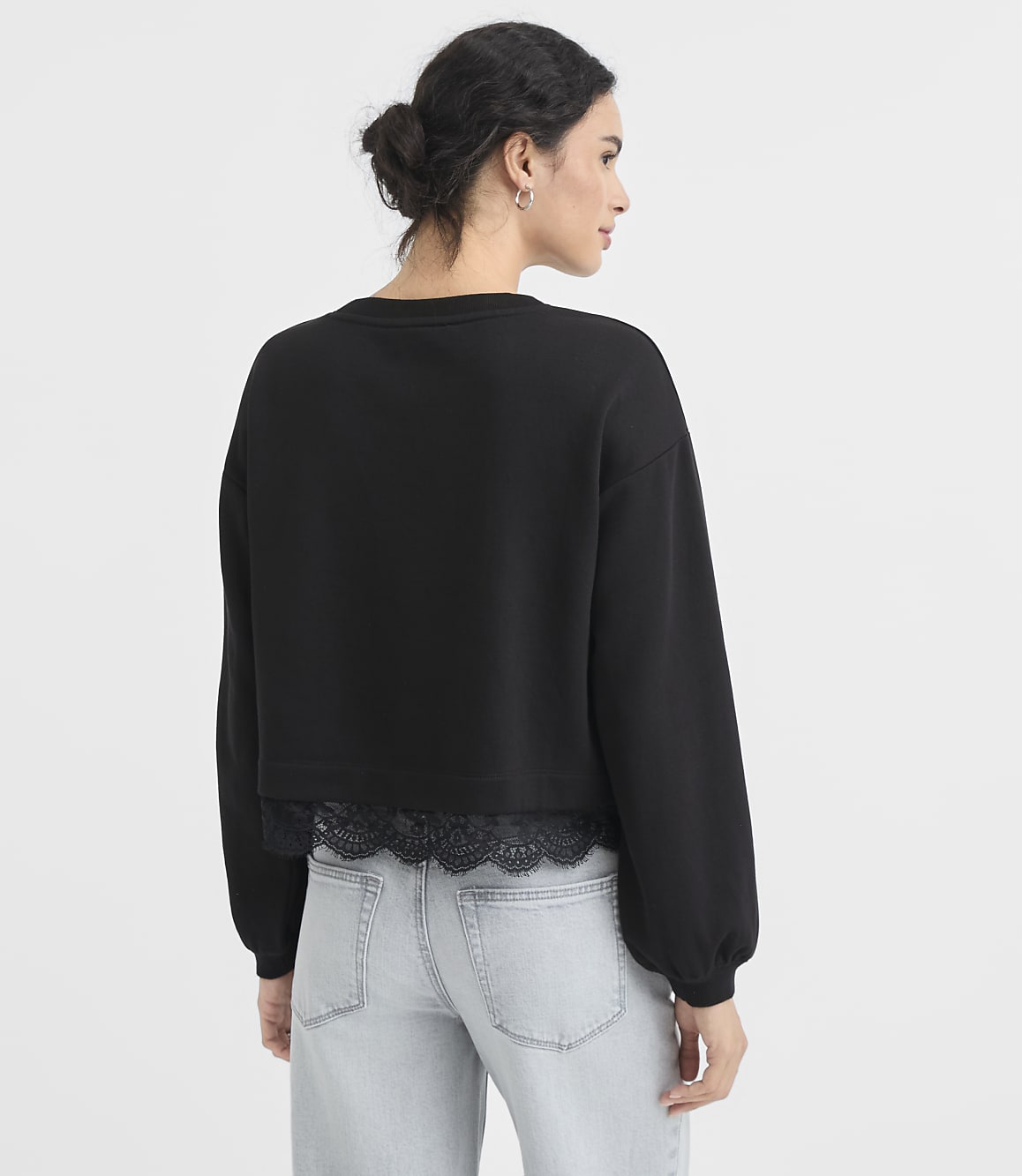 A woman in a black sweatshirt with lace trim and light wash jeans stands against a plain white background.