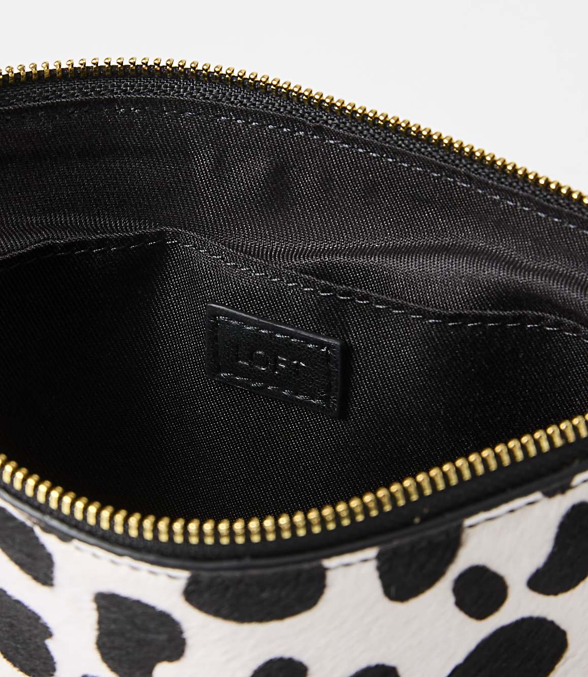 A black and white spotted clutch with a gold zipper is open to reveal its black interior, which has a "LOFT" logo tag.