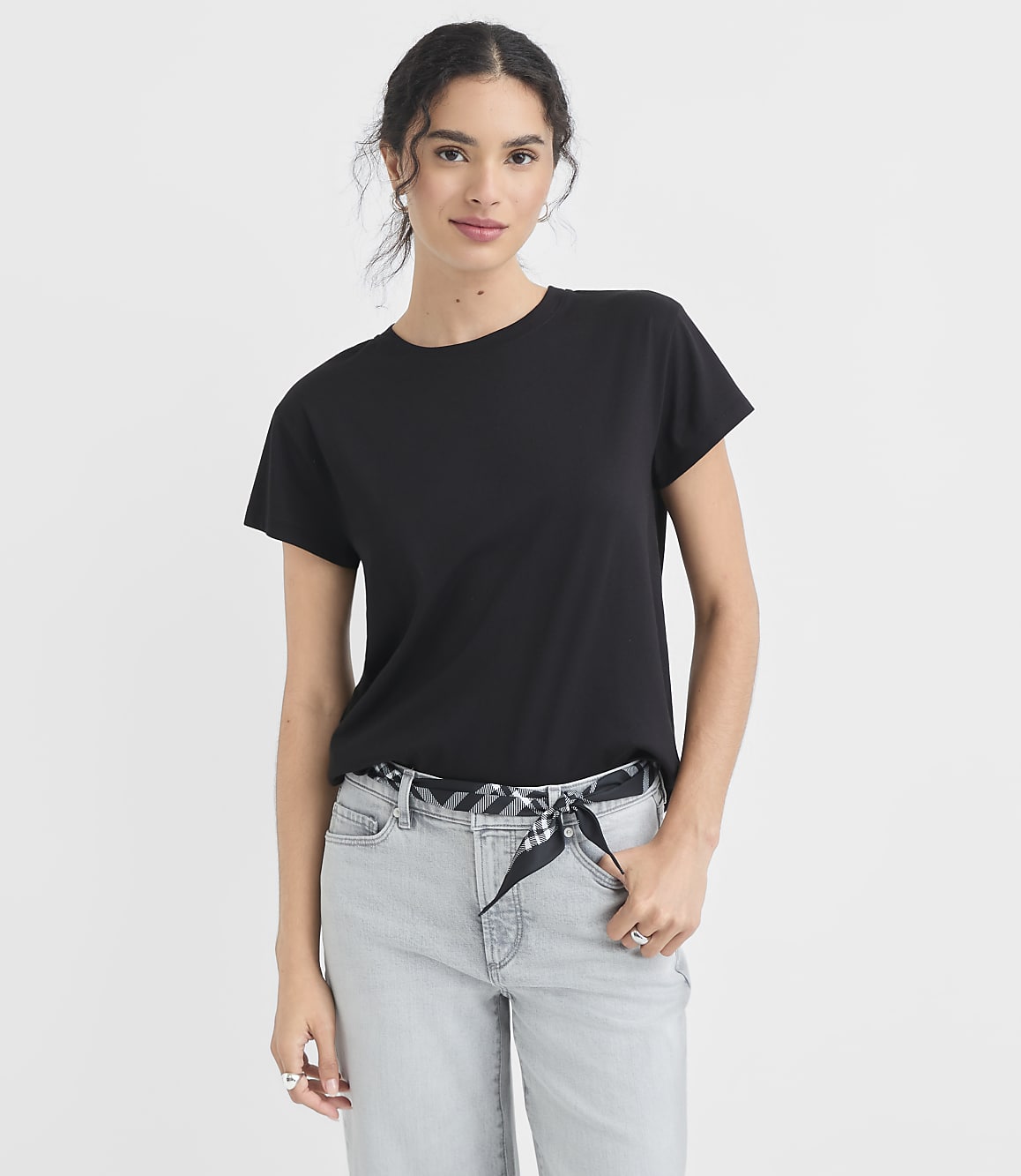 A woman in a black t-shirt and light-wash jeans stands against a plain white background.