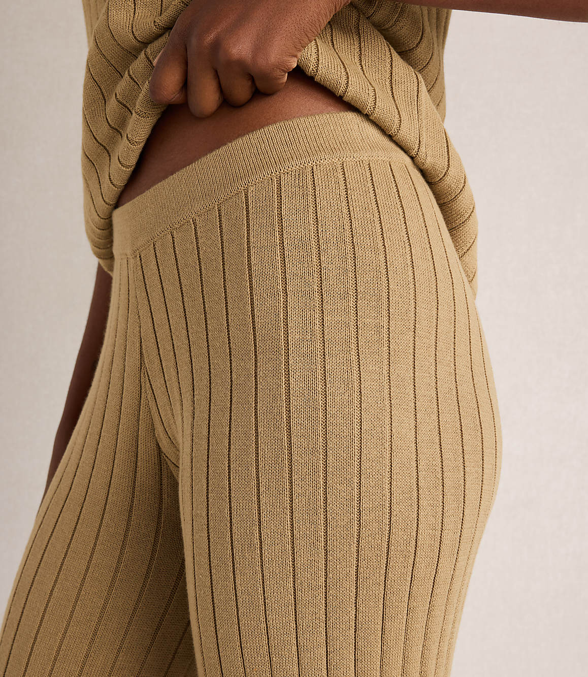 A close-up of a person wearing ribbed knit beige pants and a matching top, with a plain background.