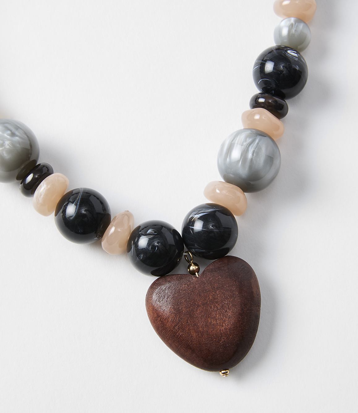 A wooden heart pendant hangs from a necklace of black, gray, and peach beads against a plain white background.