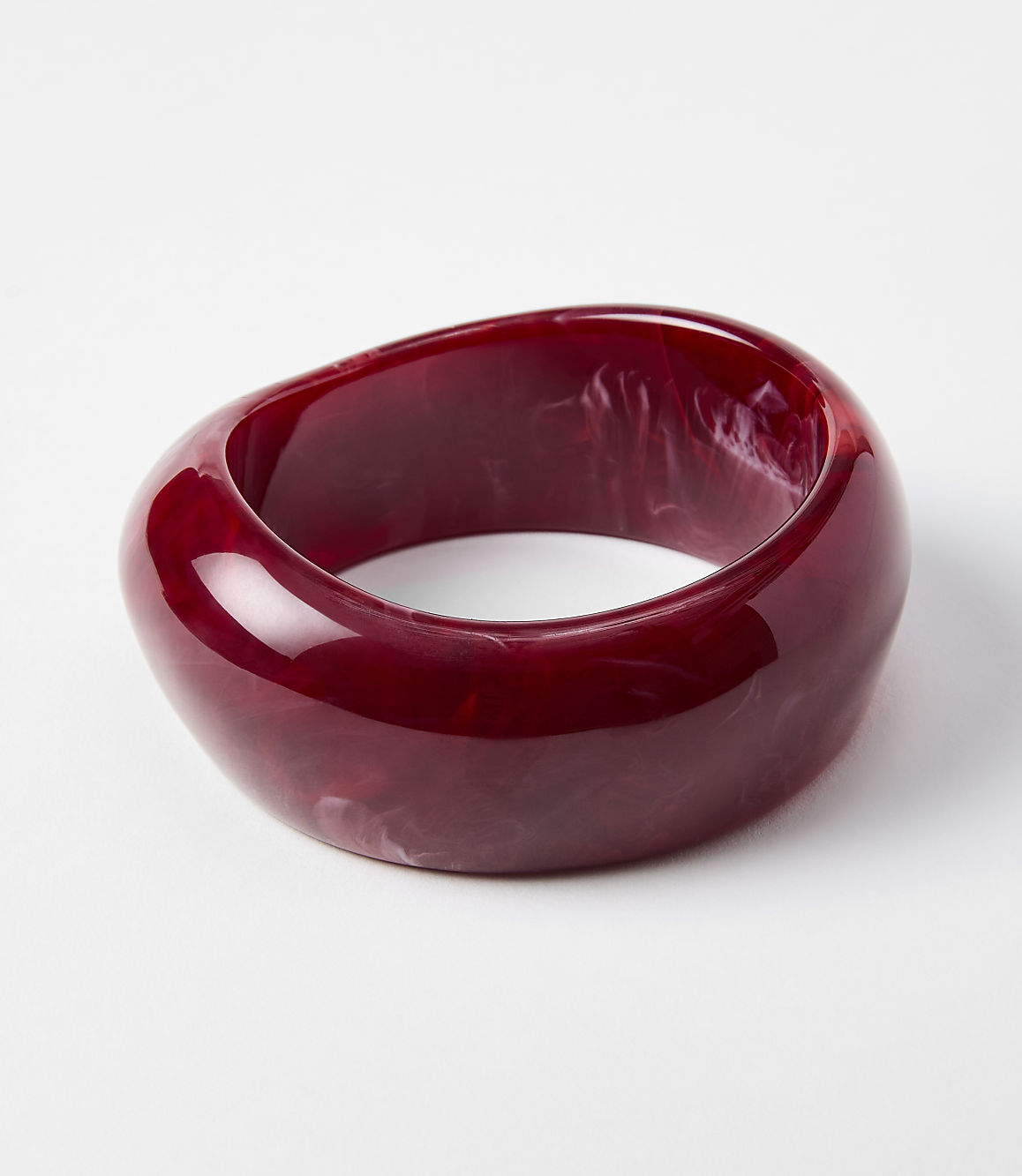 A glossy, deep red, abstractly shaped bangle with swirling white inclusions rests on a plain white background.