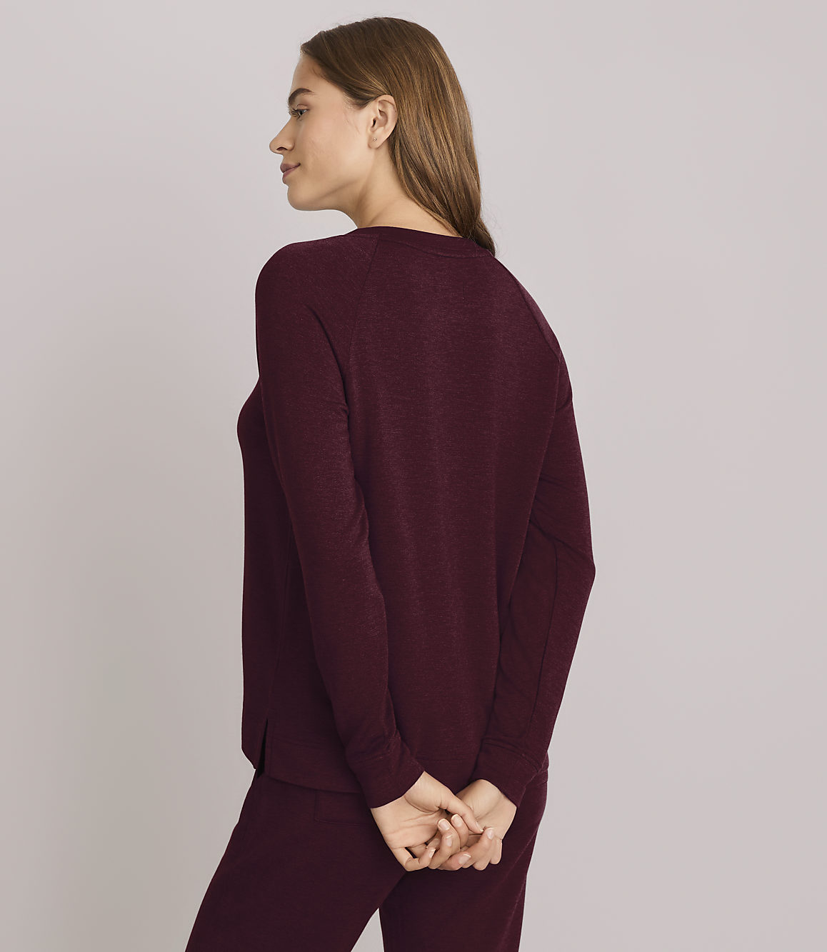 A woman in a purple sweatshirt and pants stands with her hands clasped behind her back against a plain gray background.