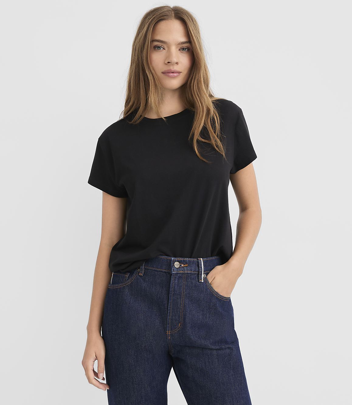 A woman in a black t-shirt and dark denim jeans stands against a plain white background.