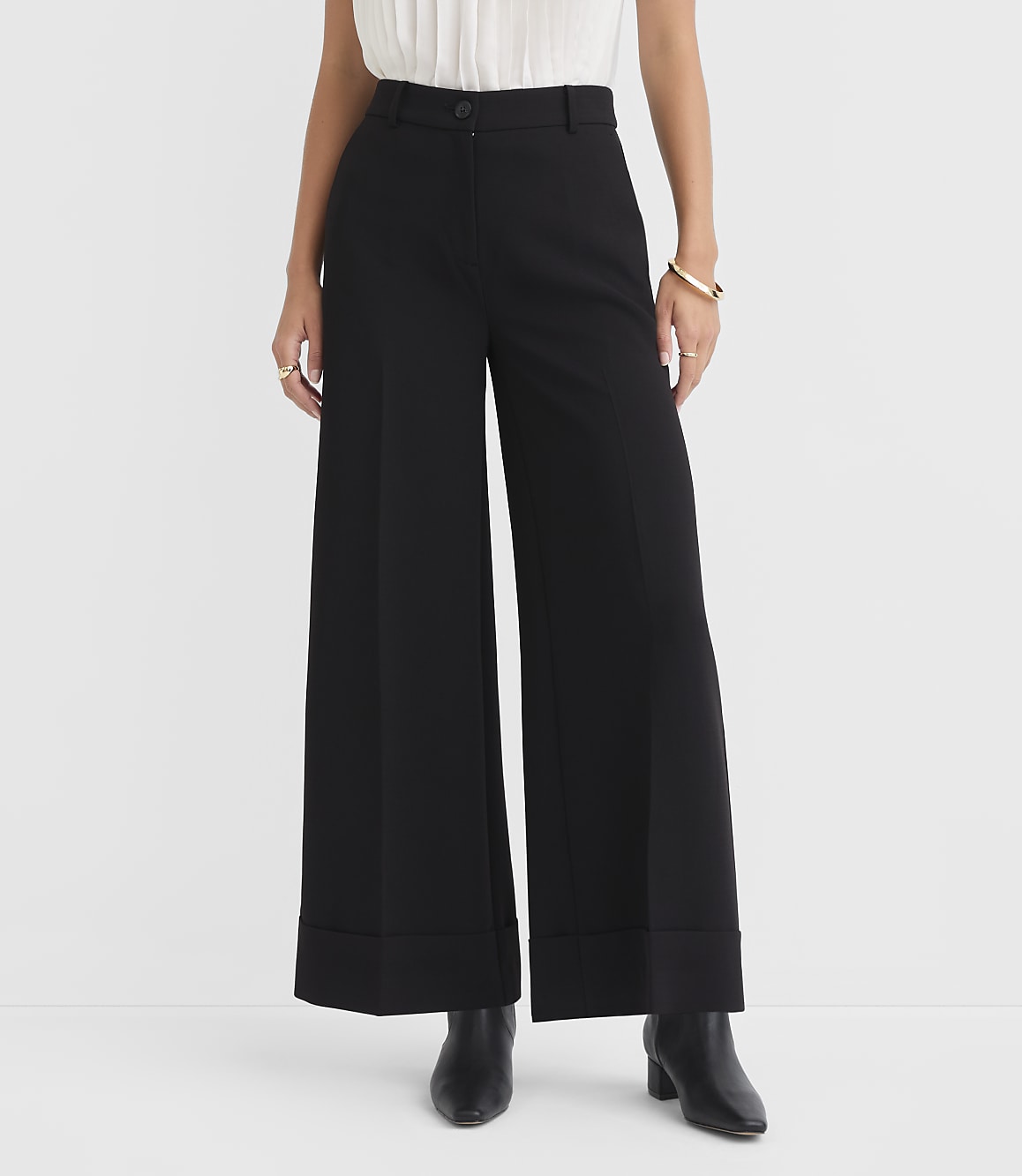 A person stands in black wide-leg pants with cuffed hems and black boots in the foreground, against a plain white background.
