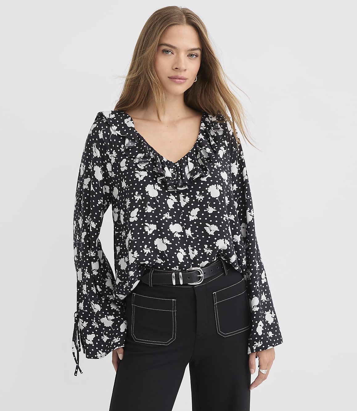 A woman in a black and white floral blouse with bell sleeves stands against a plain white background.