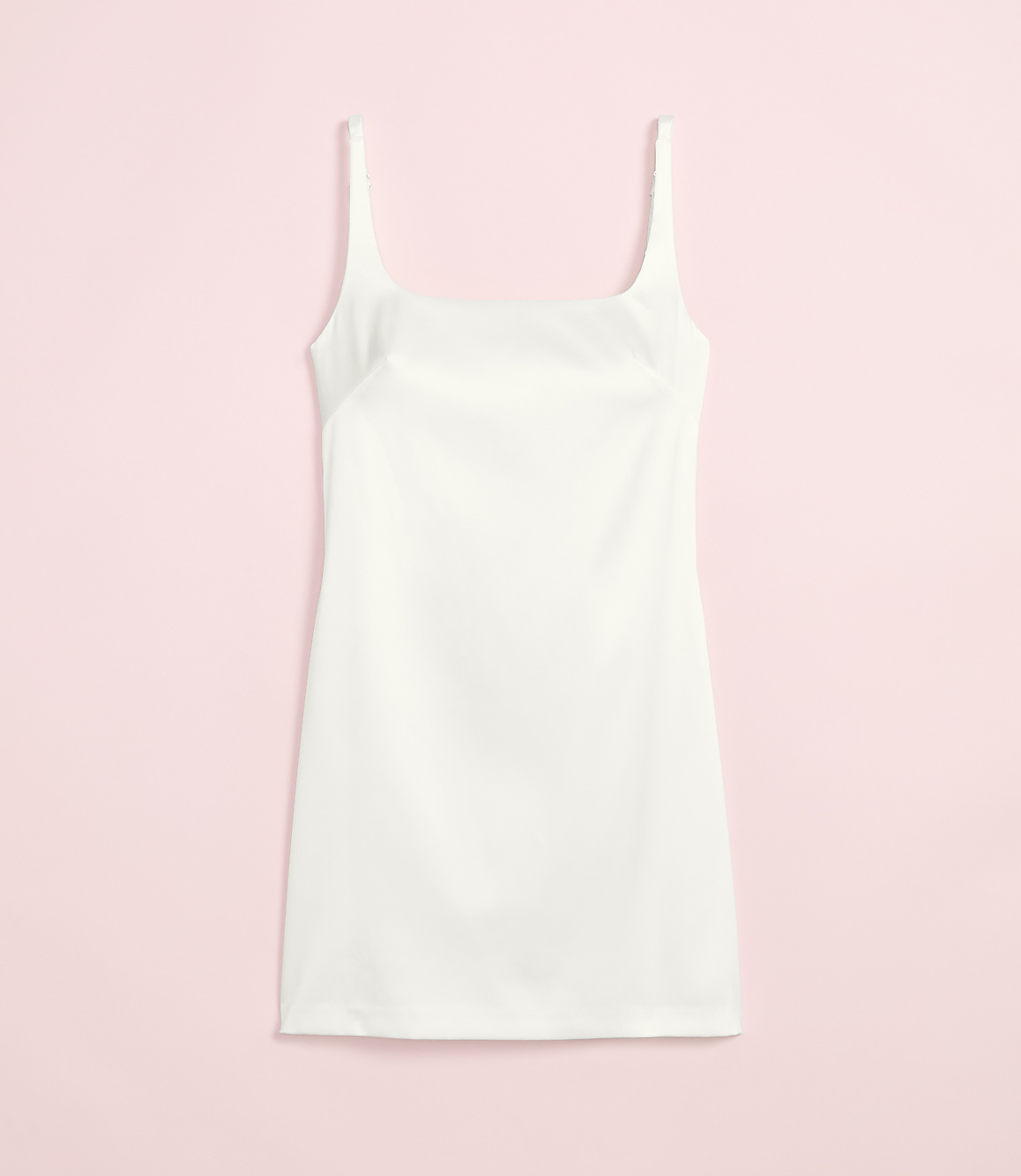 A white slip dress with thin straps is laid flat against a soft pink background.