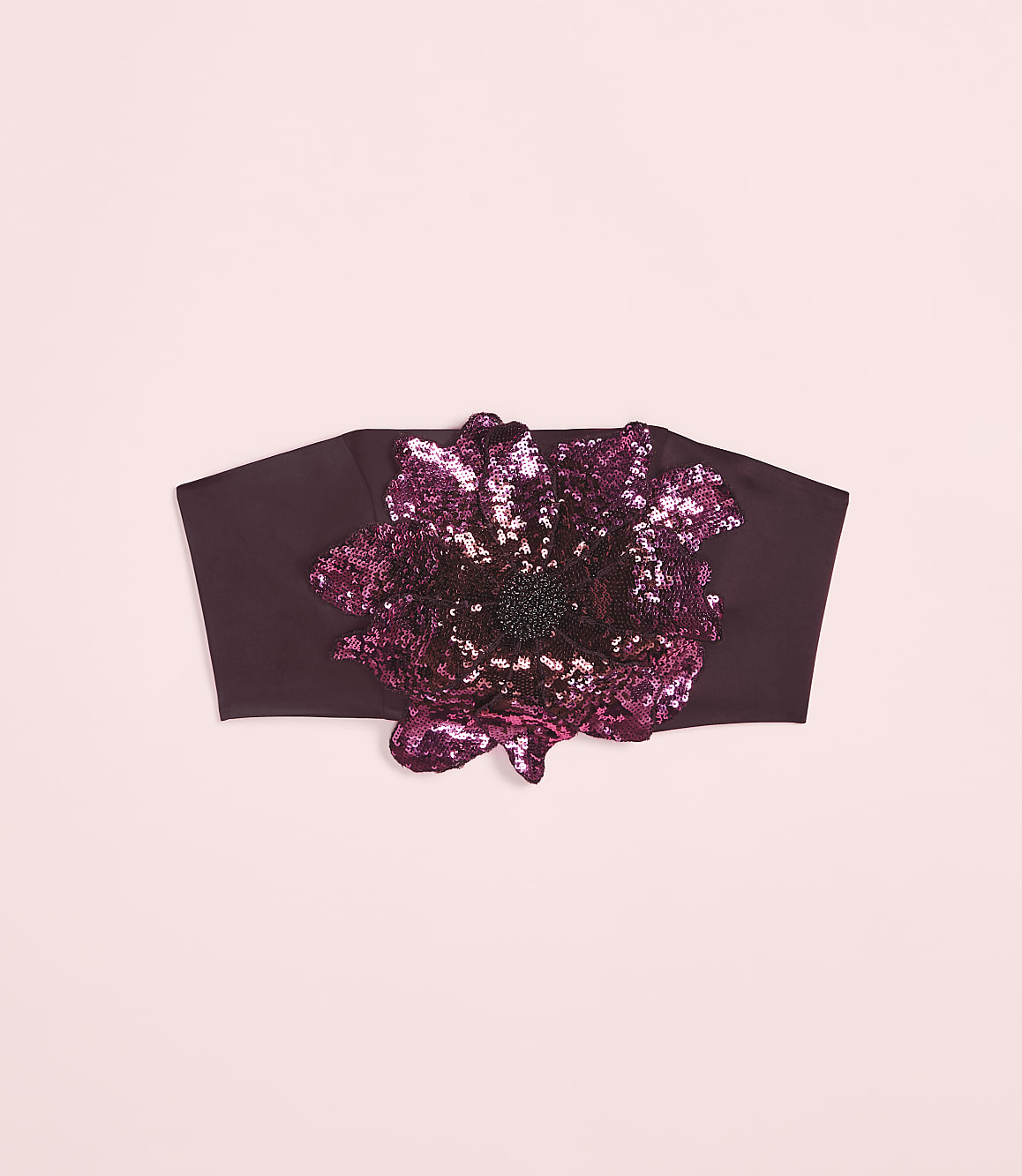 A dark plum headband with a large, sequined floral embellishment sits against a soft pink background.