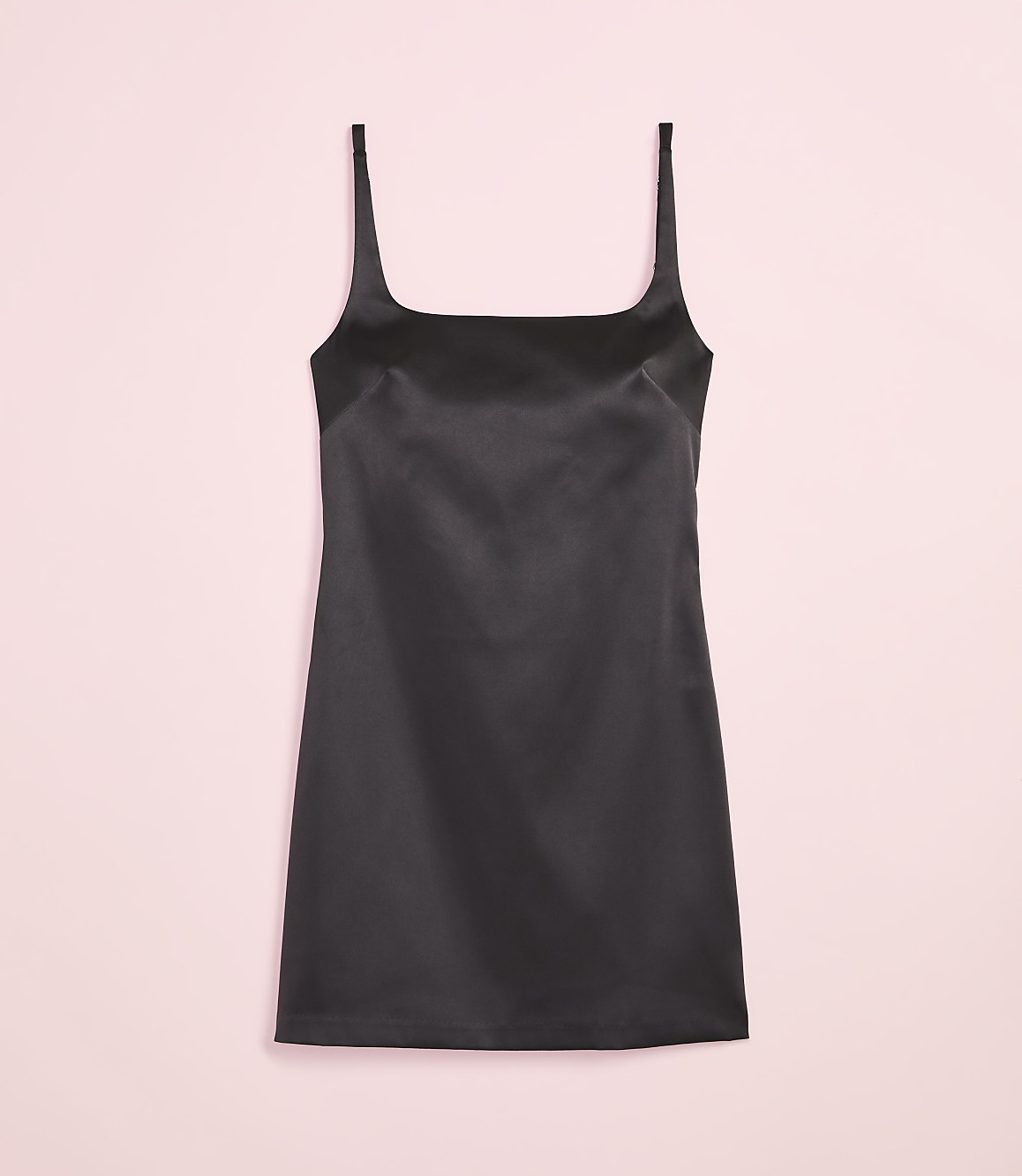 A black satin slip dress with thin straps is centered against a soft pink background.