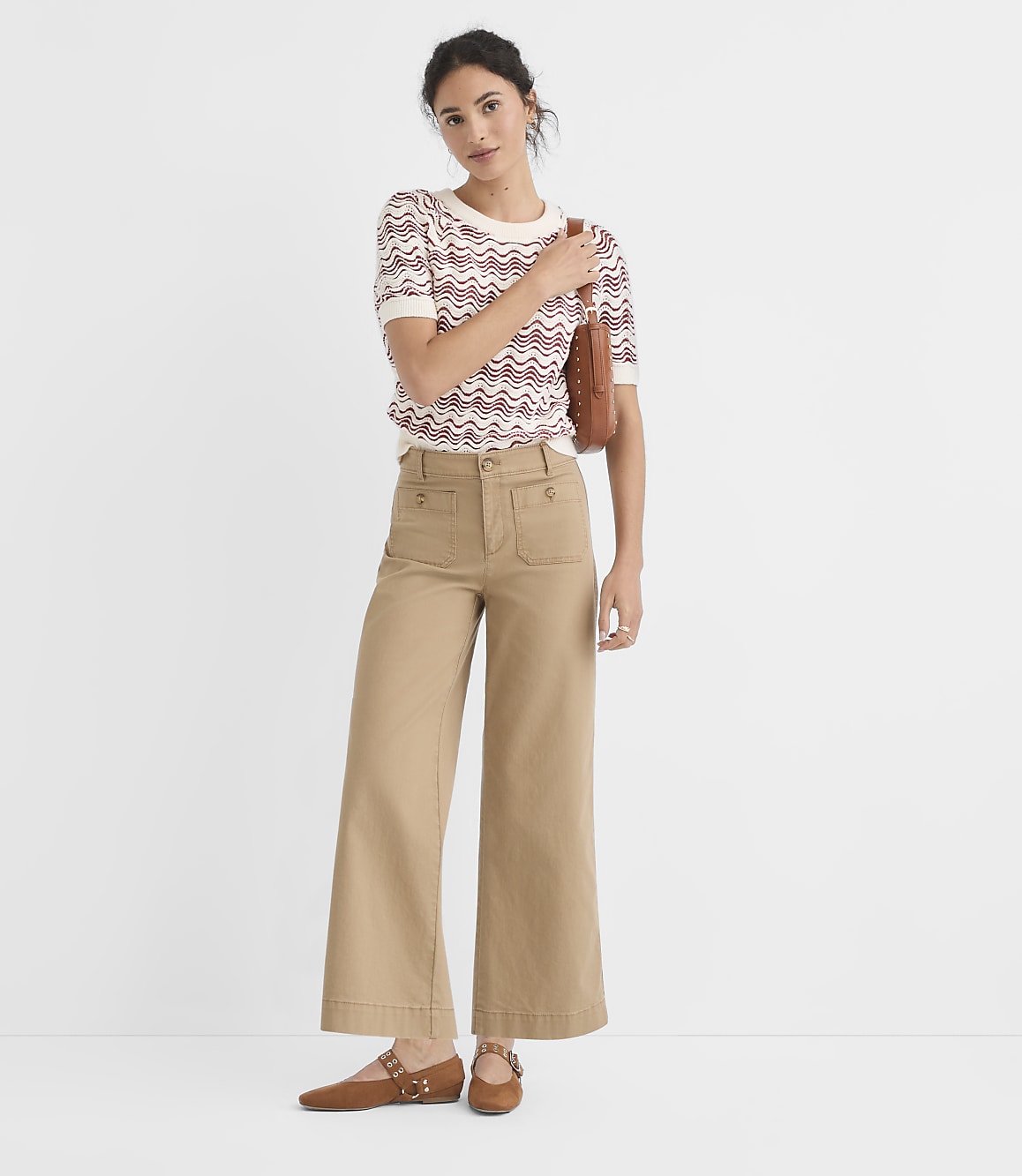 A woman in a patterned sweater and tan pants stands against a plain white background.