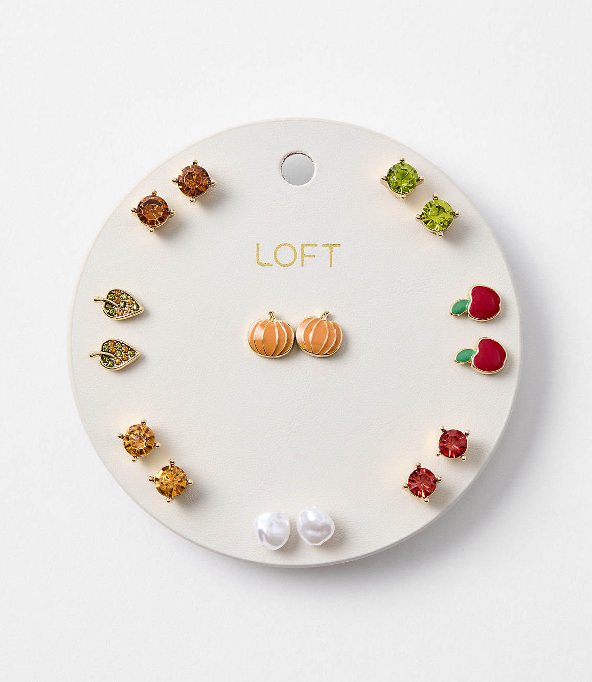 A circular white card displays an assortment of small, colorful stud earrings, including pumpkins, leaves, apples, and gemstones, against a plain white background.