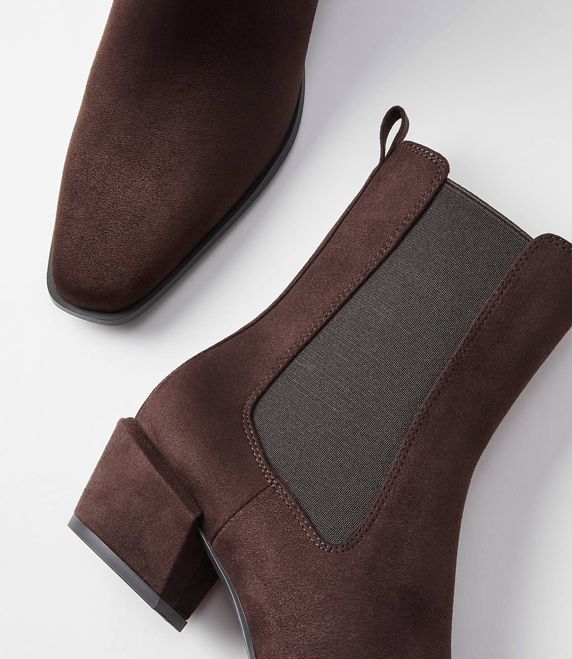 A pair of brown suede Chelsea boots with block heels are arranged on a white background, with one boot in the foreground and the other partially visible in the background.