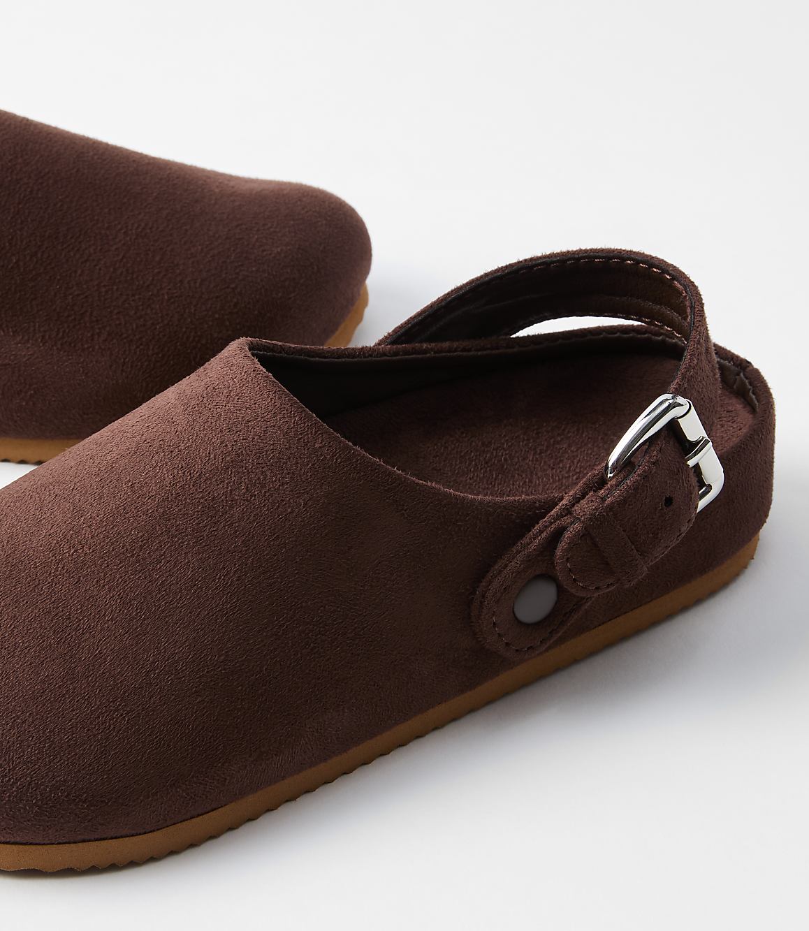 A pair of brown suede clogs with a silver buckle rests on a white surface, with another clog partially visible in the background.