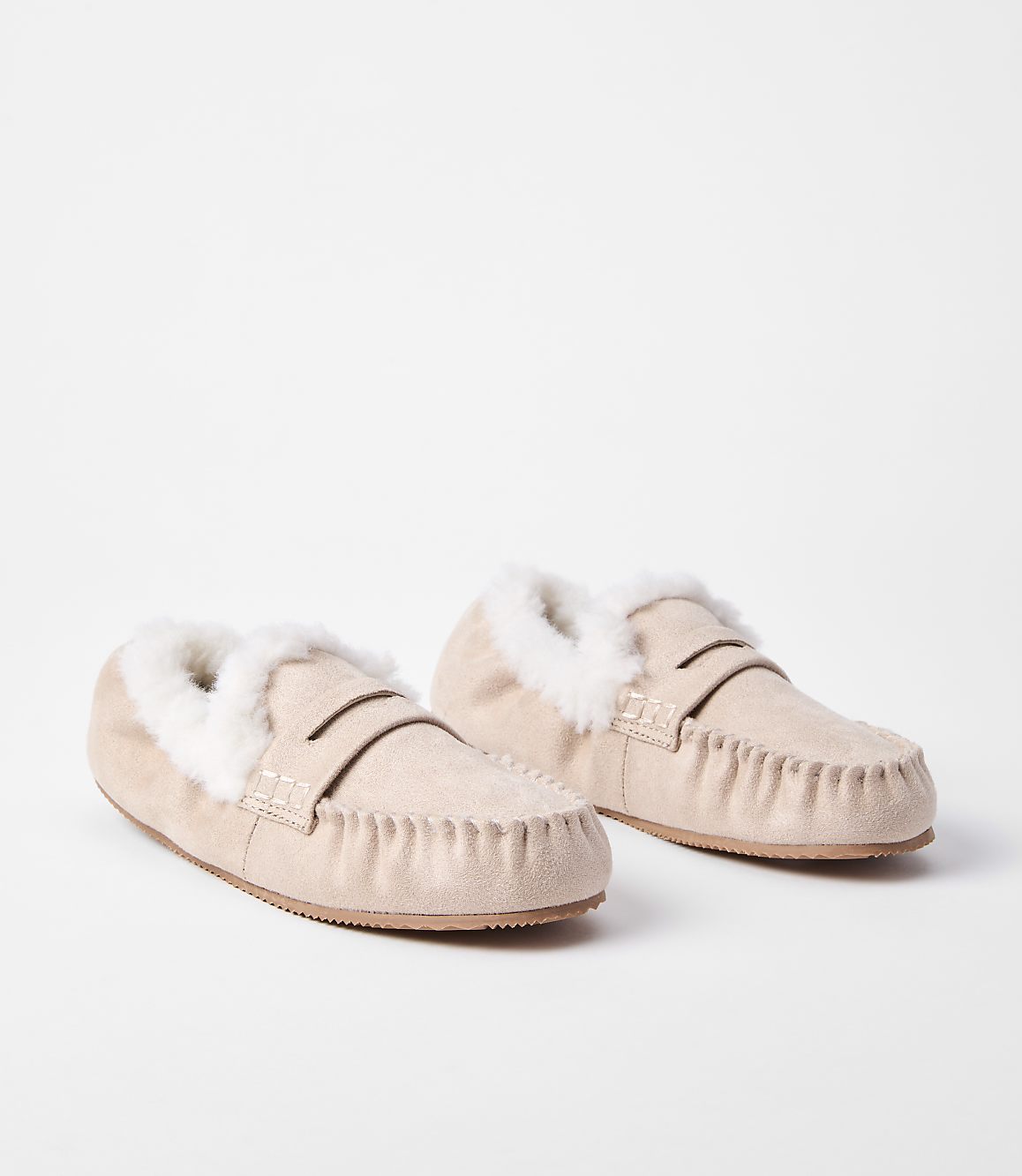 A pair of light beige moccasin slippers with fluffy white lining rests on a plain white background.