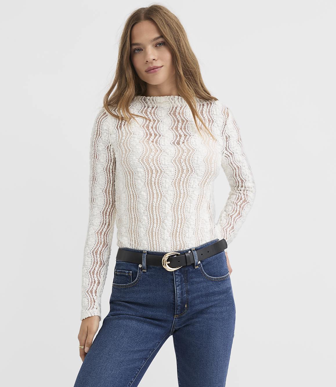 A woman in a white lace top and blue jeans stands against a plain white background.