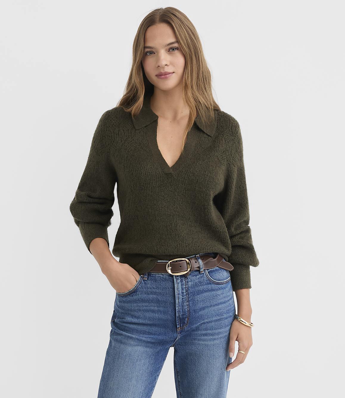 A woman in a dark green collared sweater and blue jeans stands against a plain white background.
