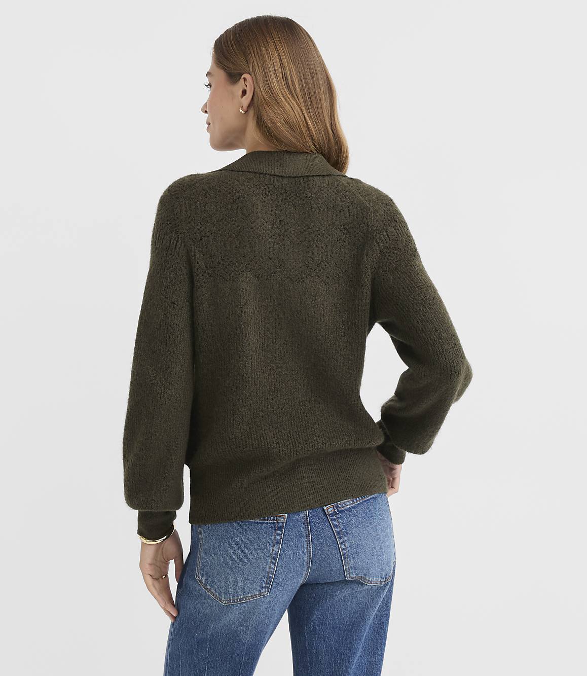 A woman in a textured olive green sweater and blue jeans stands against a plain, light gray background.