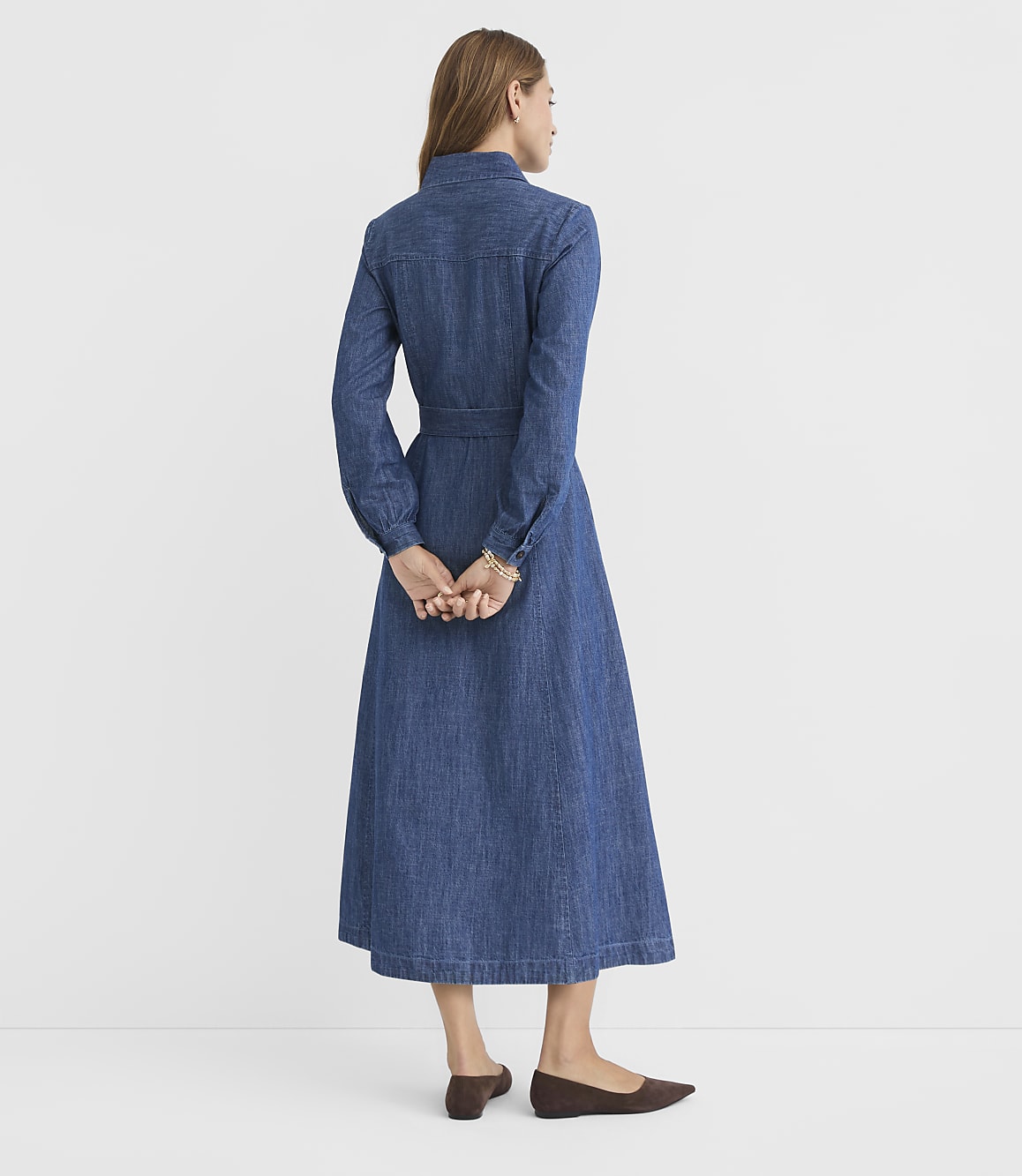 A woman in a denim dress stands with her hands clasped behind her back against a plain white background.