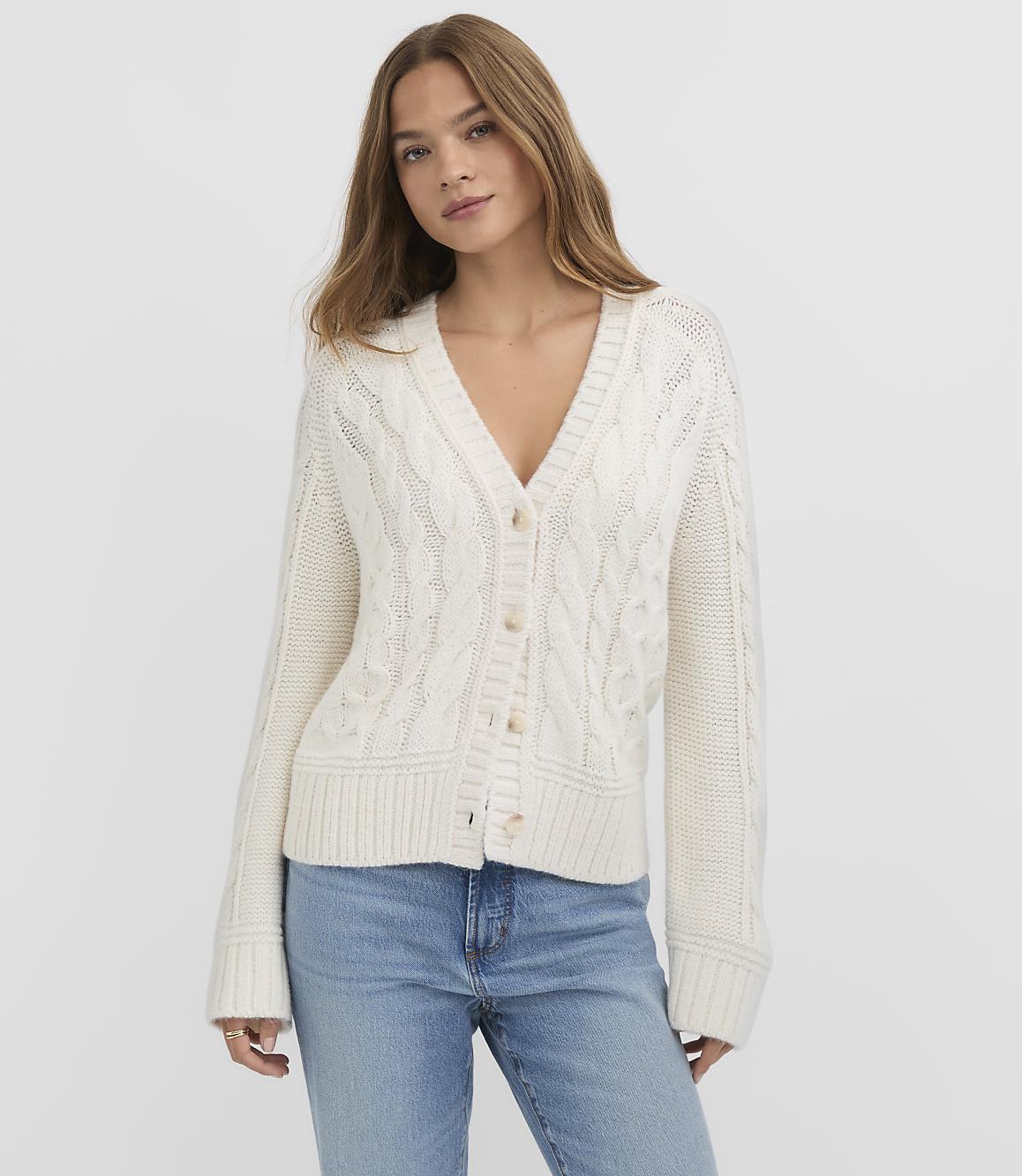 A woman in a cream cable-knit cardigan and blue jeans stands against a plain white background.