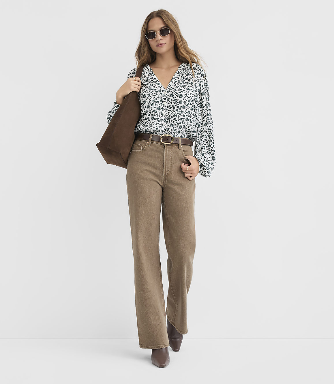 A woman in a floral blouse and wide-leg brown pants stands against a plain white background, carrying a brown tote bag.