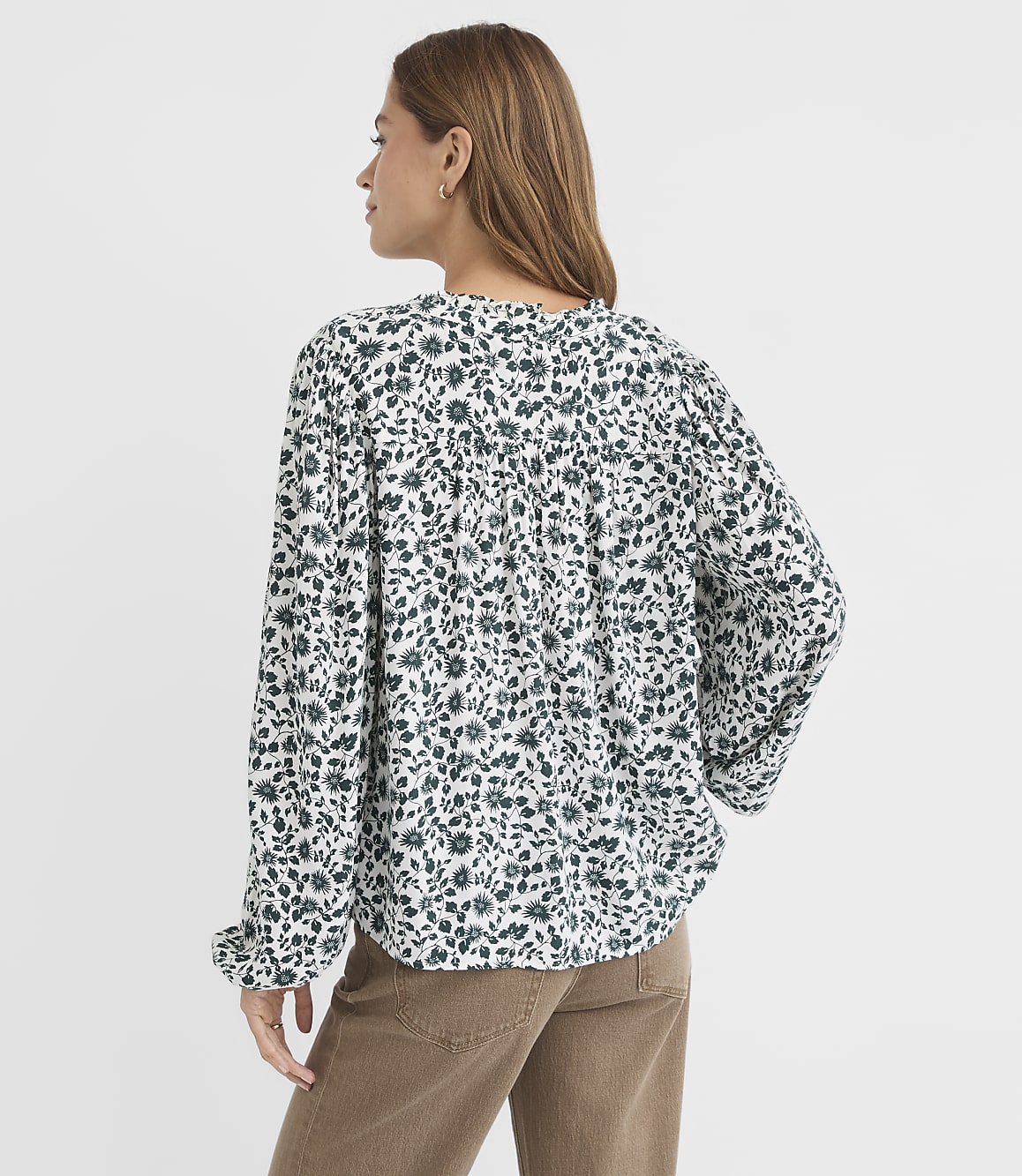 A woman in a white and green floral blouse and brown pants stands against a plain white background.