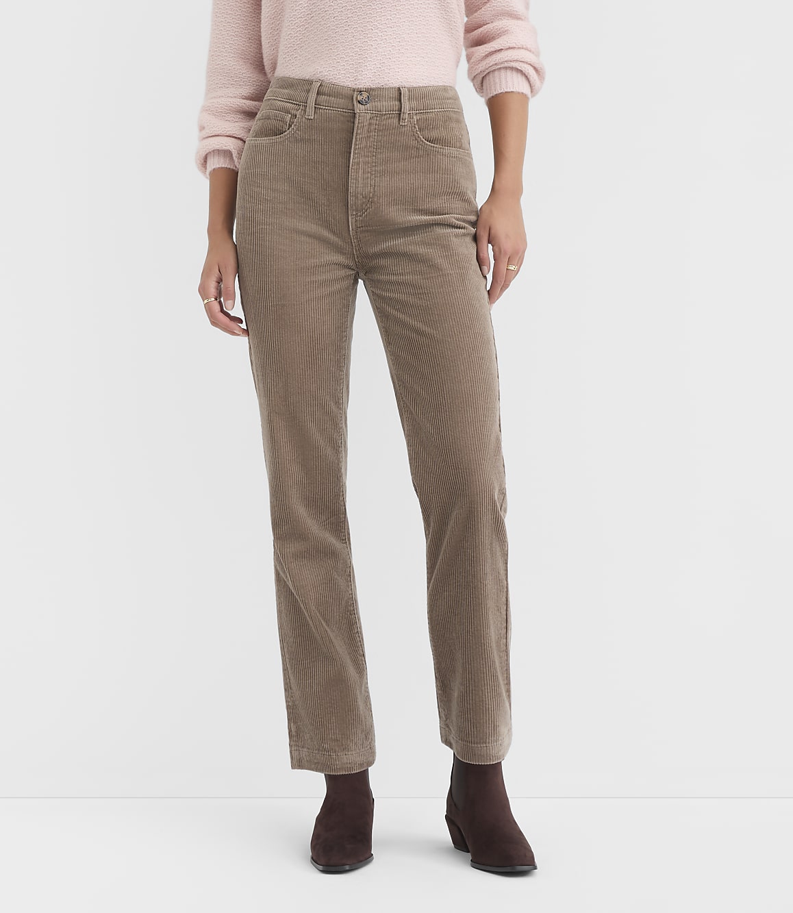 A person stands in the foreground wearing a pink sweater and tan corduroy pants, with a plain white background.