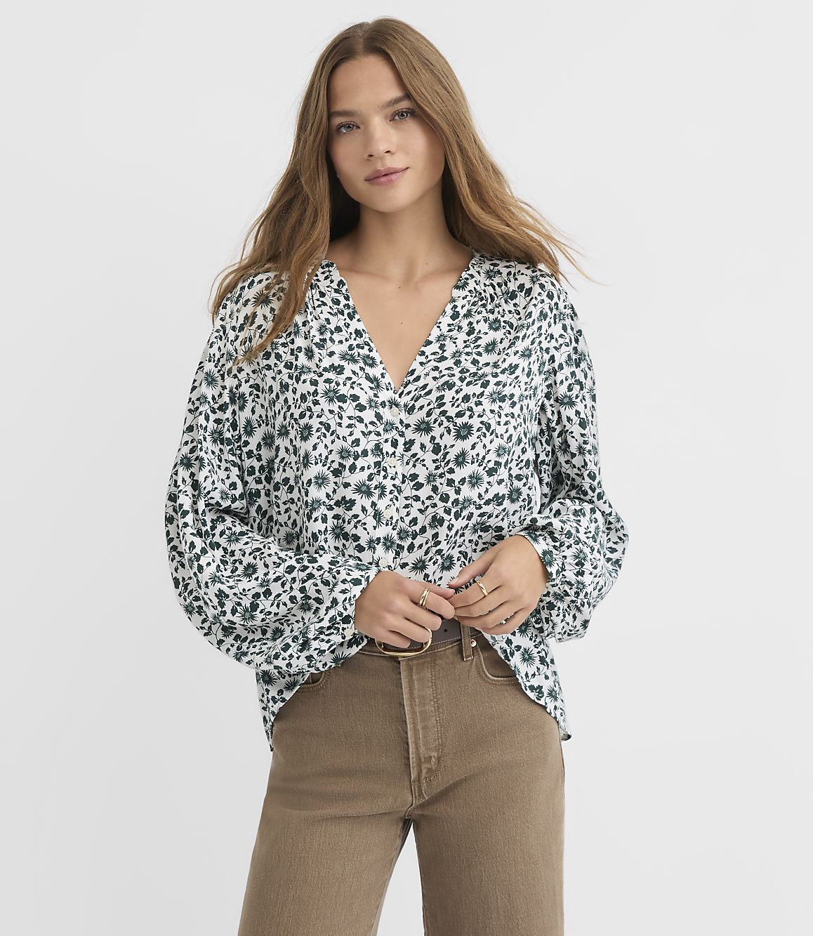 A young woman in a floral blouse and brown pants stands against a plain white background.