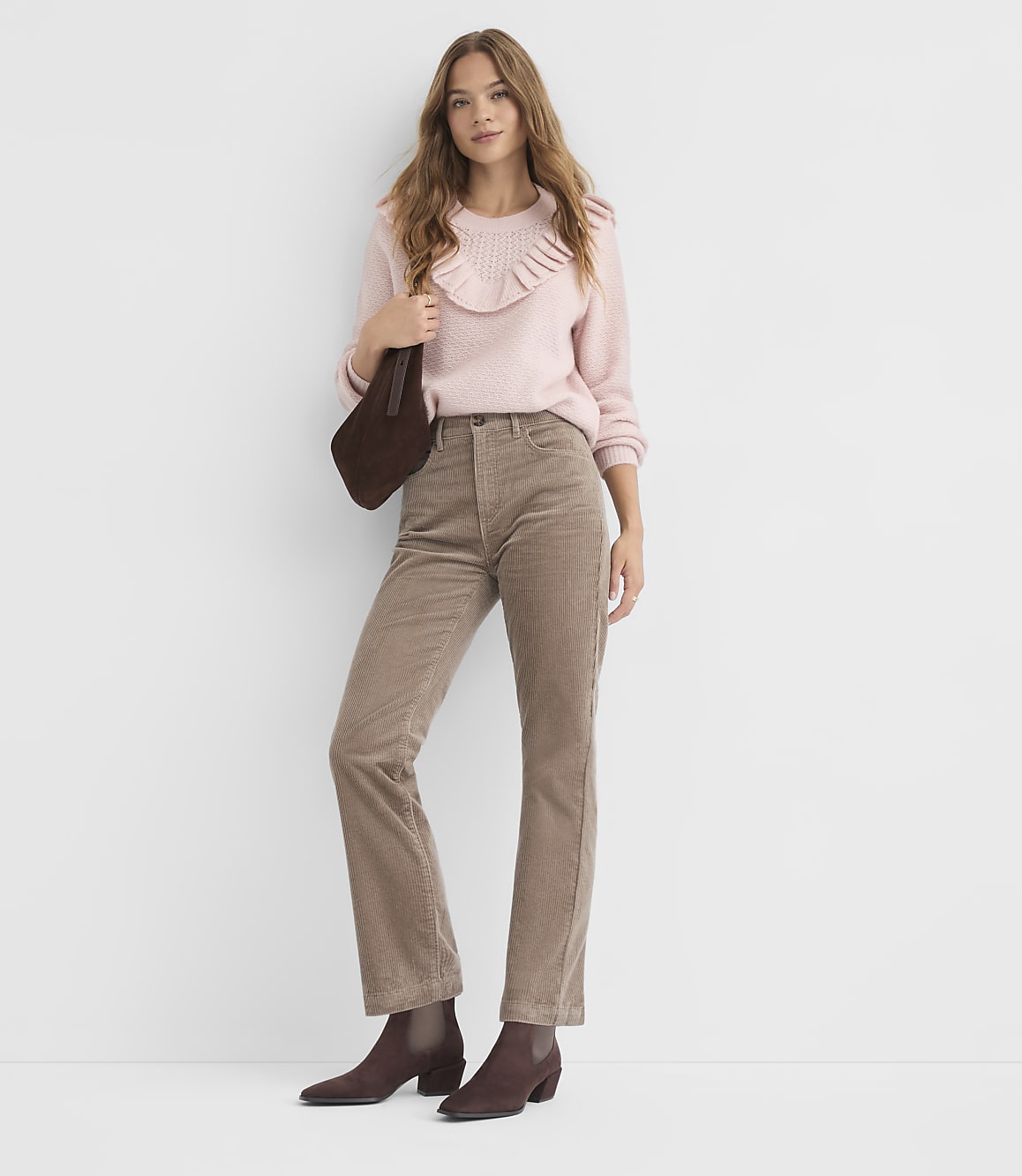 A woman in a pink ruffled sweater and brown corduroy pants stands against a plain white background, holding a dark brown purse.