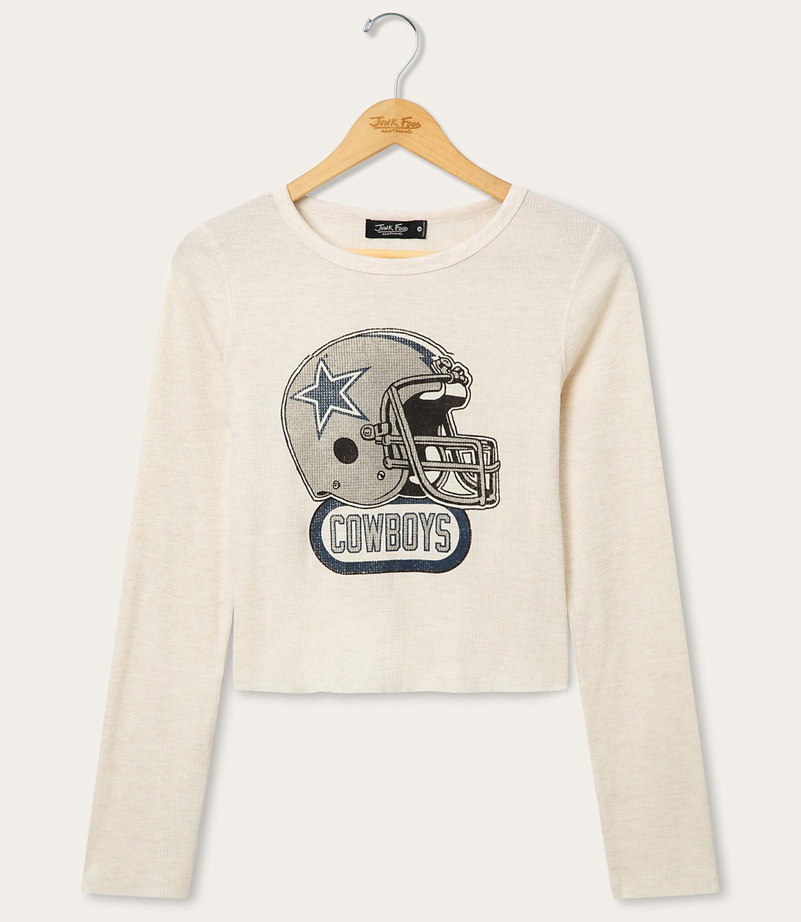 A cream-colored long-sleeved shirt with a graphic of a Dallas Cowboys helmet is displayed on a hanger against a plain white background.