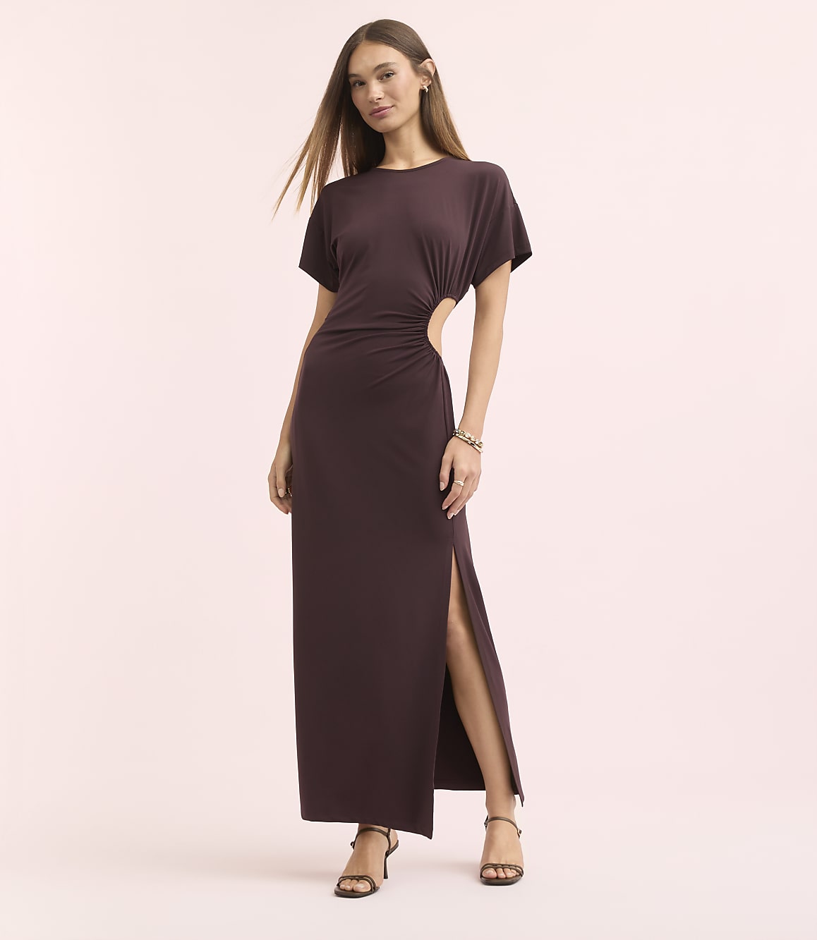 A woman in a dark purple, cutout maxi dress stands against a soft pink background.