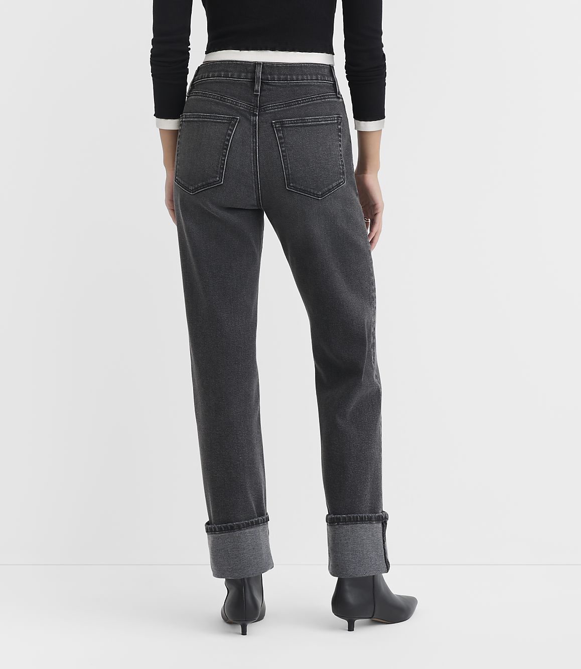 A person wearing dark wash cuffed jeans and black heeled boots stands against a plain white background.