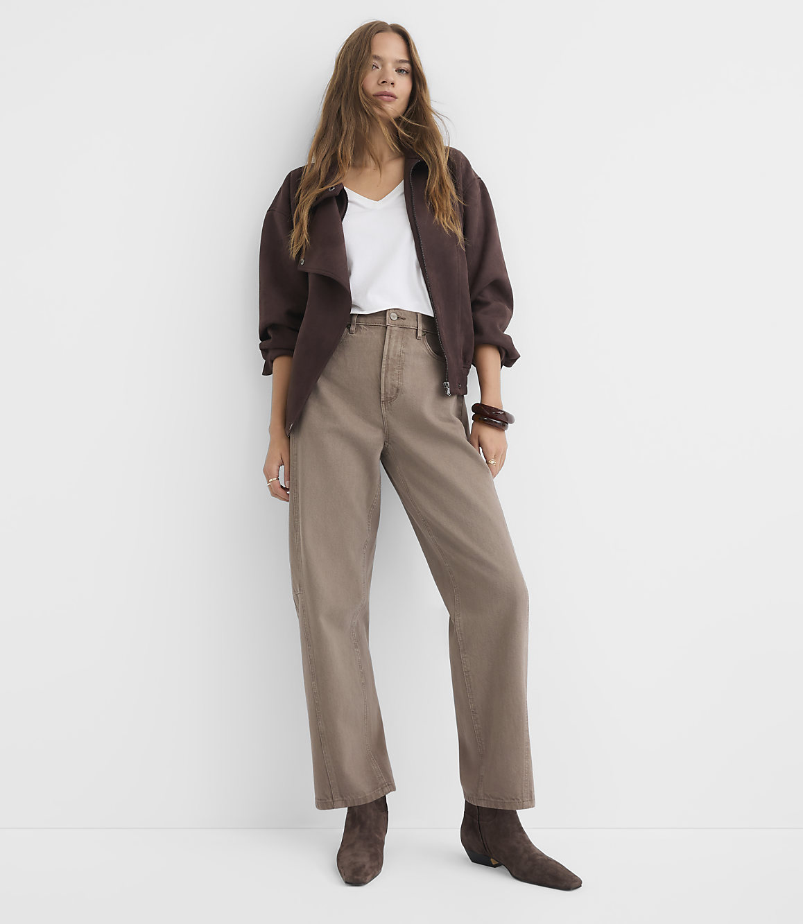 A woman in a brown jacket and taupe pants stands against a plain white background.