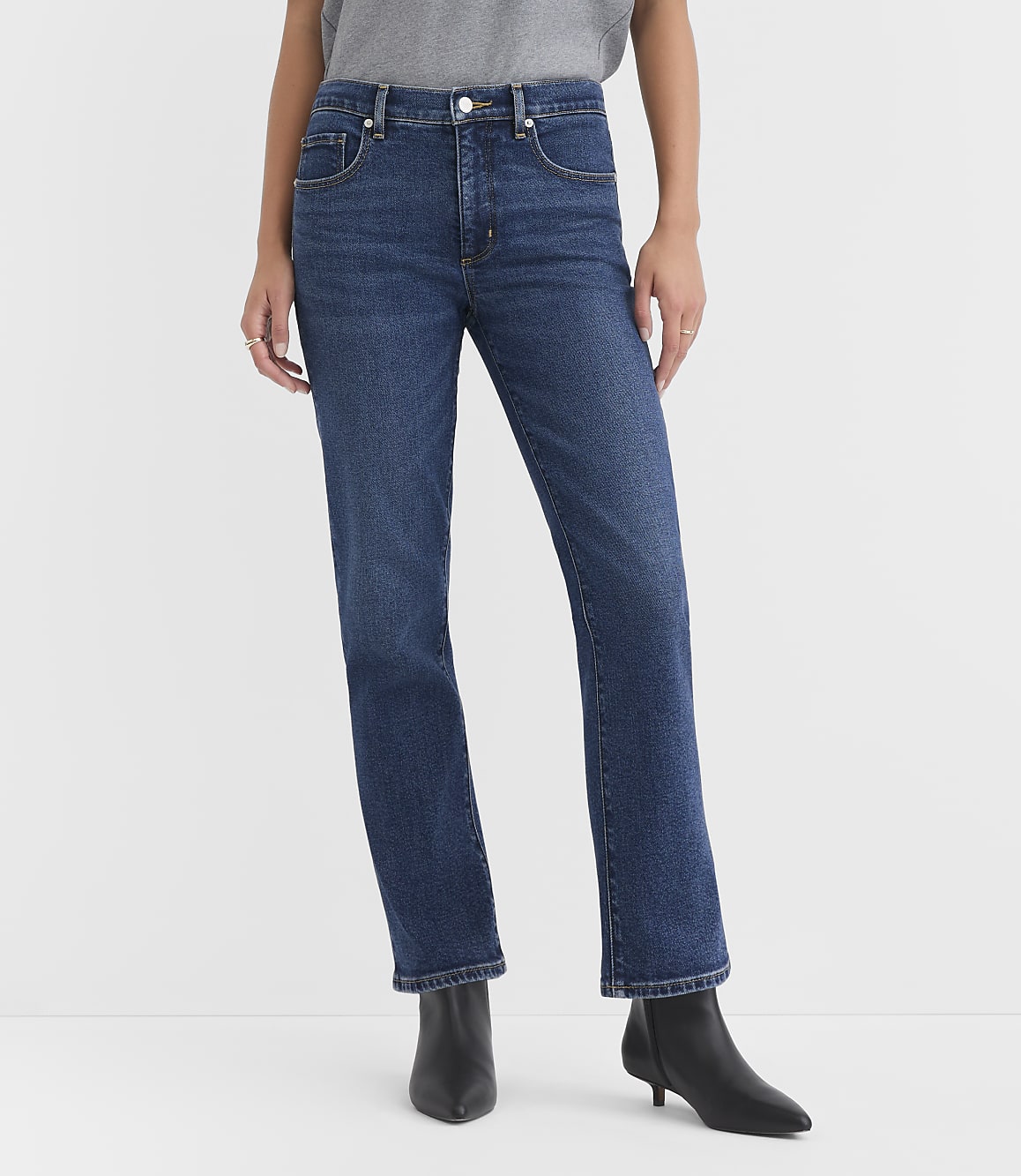 A person wearing dark wash straight-leg jeans and black heeled boots stands against a plain white background.