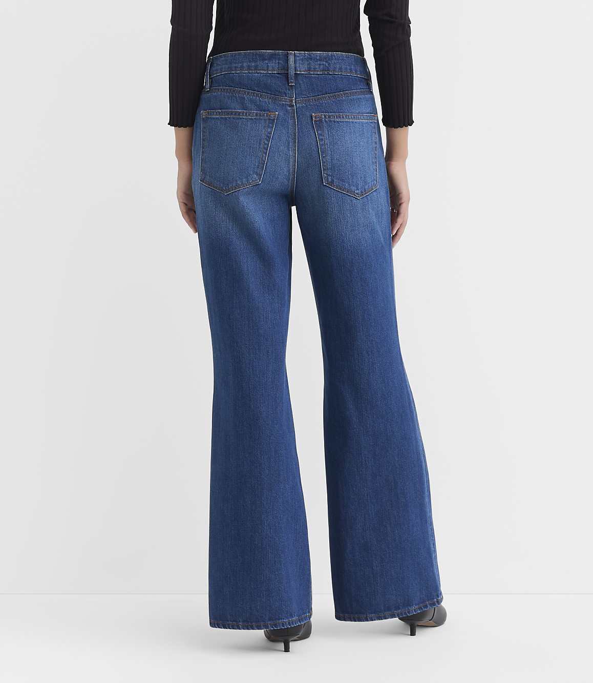A person wearing flared blue jeans and a black top stands against a plain white background.