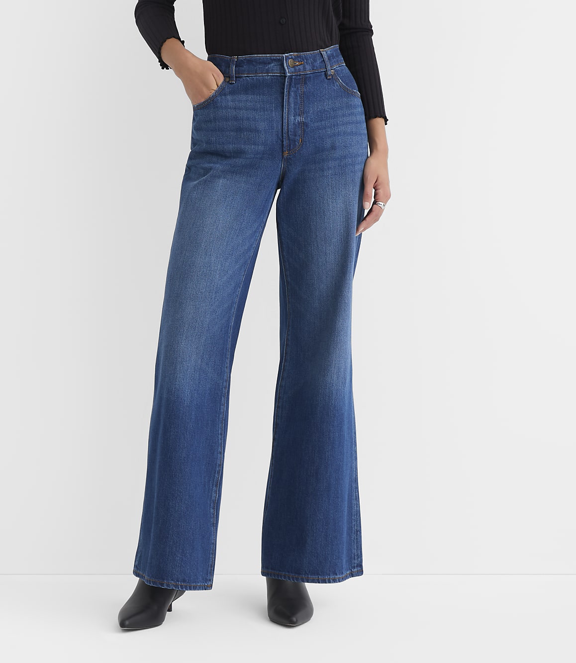 A person wearing wide-leg blue jeans and black heeled boots stands against a plain white background.