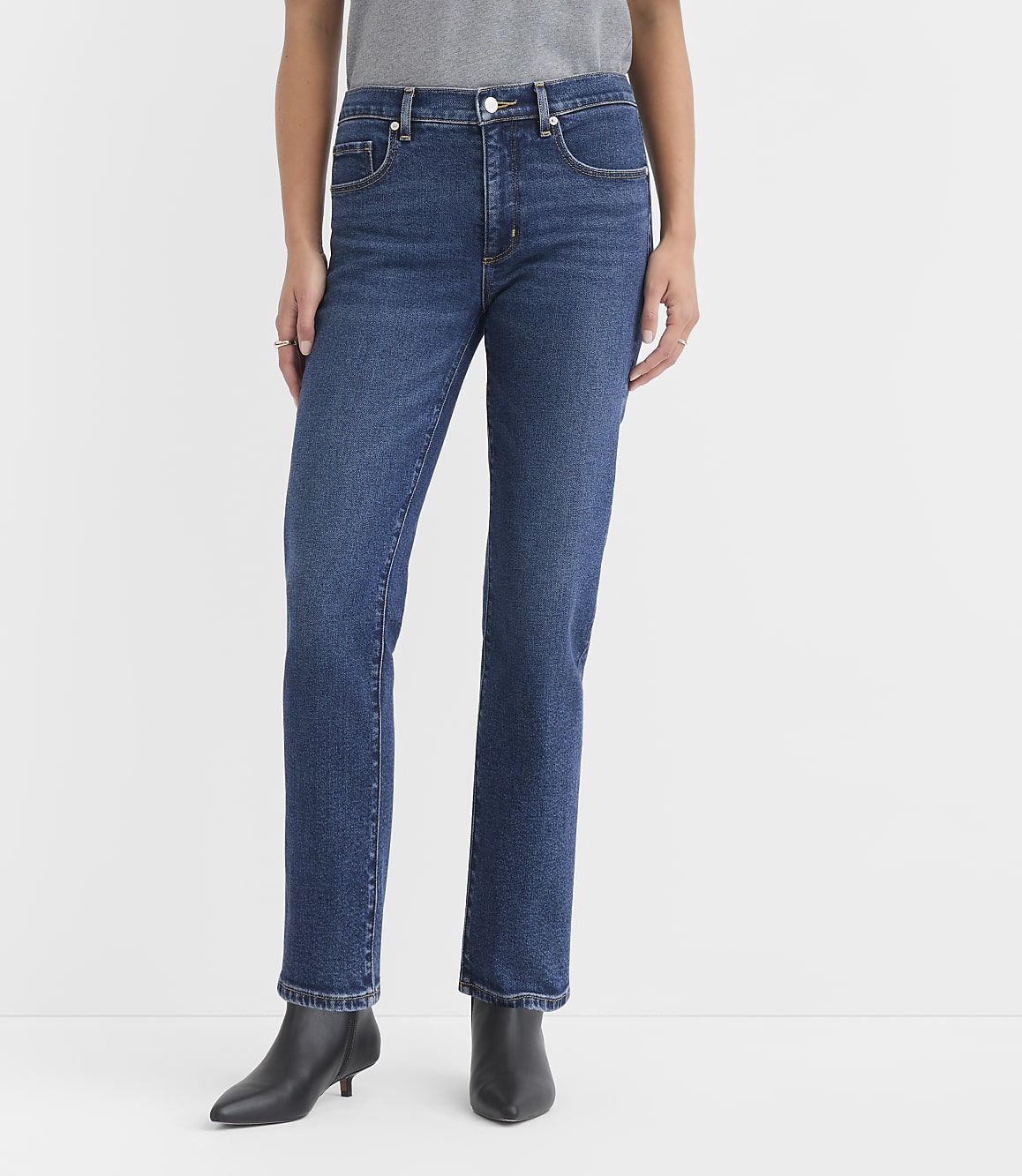 A person stands in dark wash straight-leg jeans and black ankle boots against a plain white background.