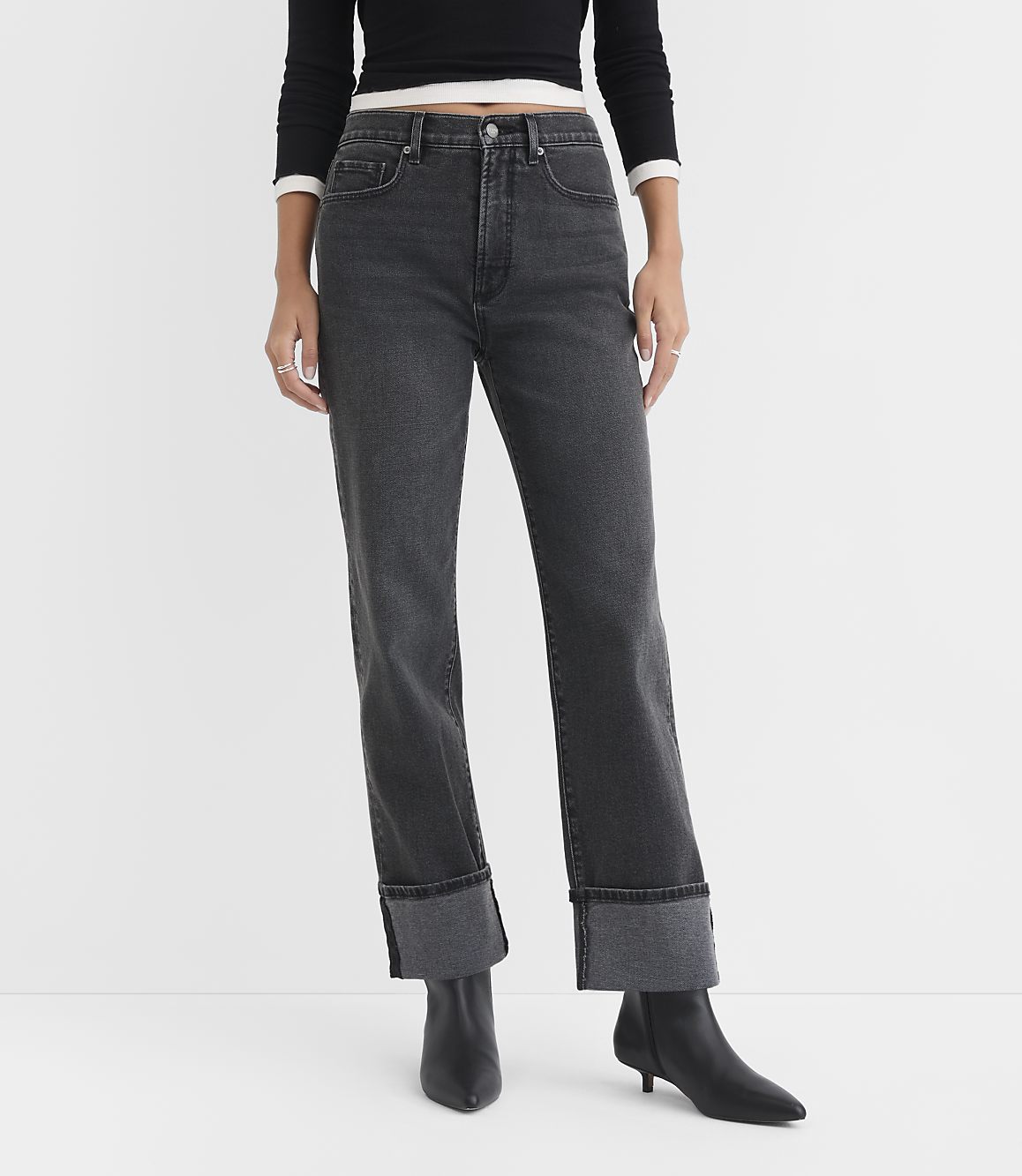 A person stands in the foreground wearing cuffed dark wash jeans and black boots against a plain white background.